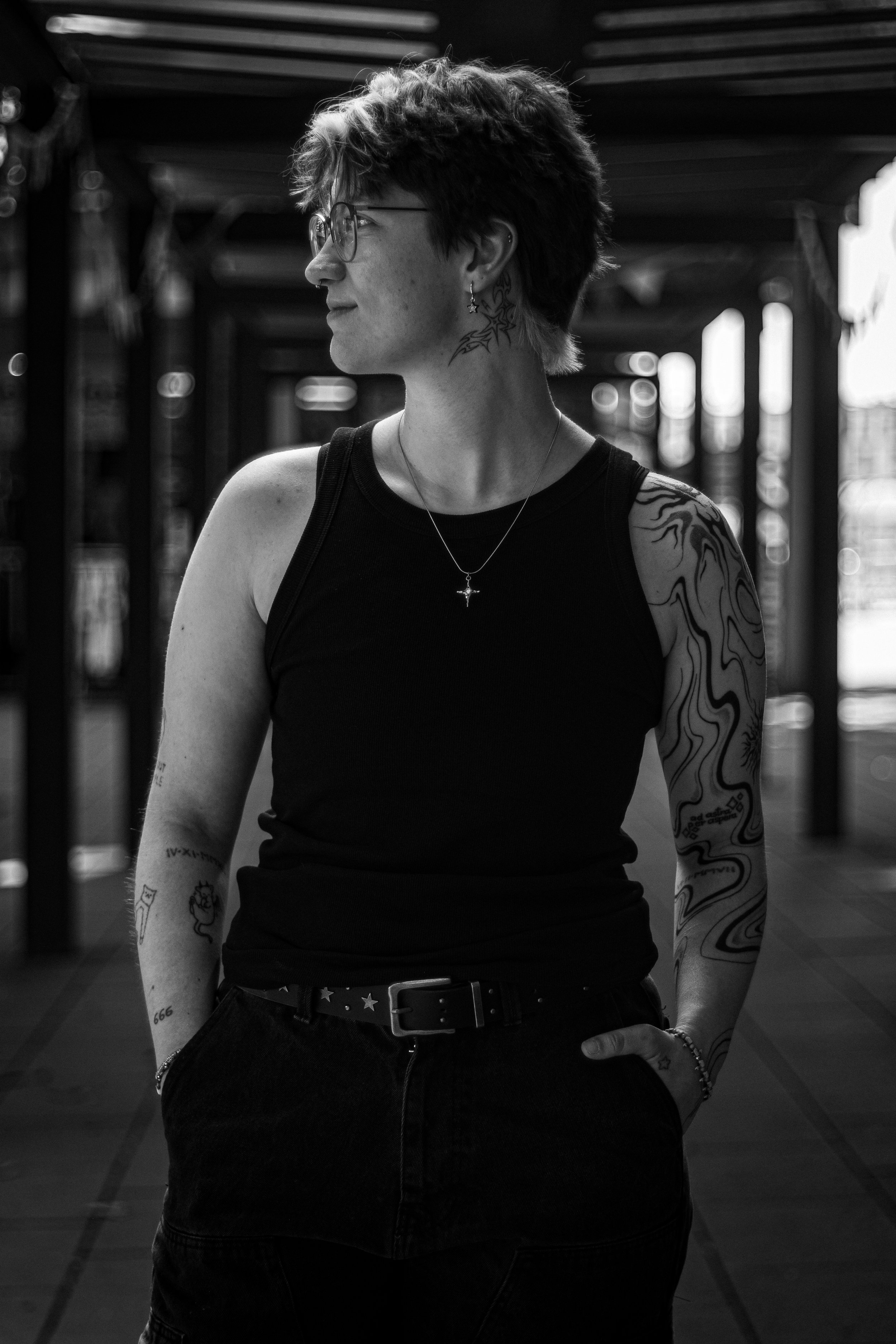 Schwarz-weiß-Foto eines jungen Menschen mit Kurzhaarfrisur, Tattoos am Arm und Hals, trägt eine schwarze Weste, eine Halskette, eine Armbanduhr und eine Ring. Er trägt Brille und steht in einem überdachten Bereich mit Holzdecke.