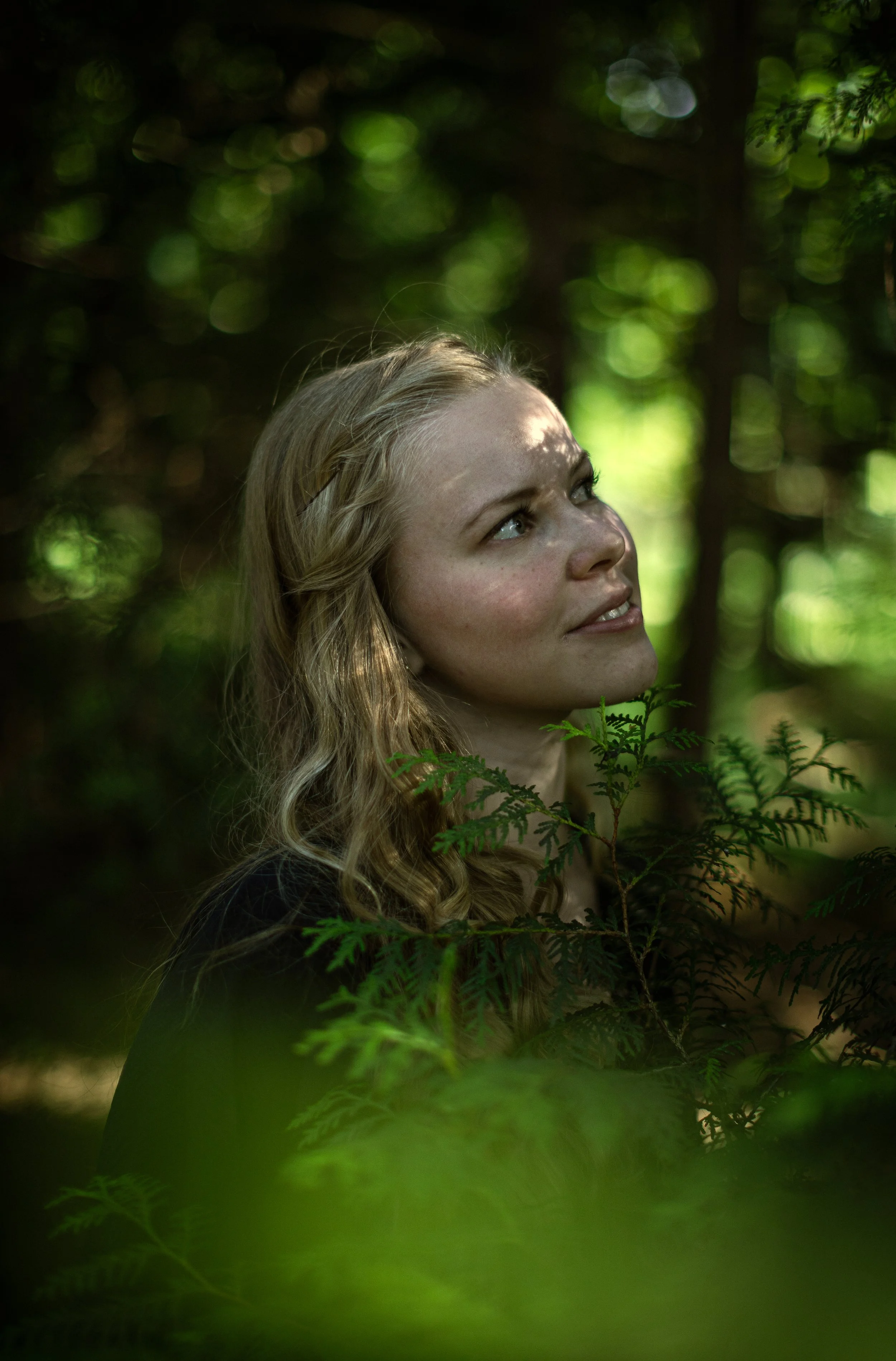 Junge Frau mit blonden, lockigen Haaren, blickt nach oben, umgeben von grünen Blättern in einem Wald.