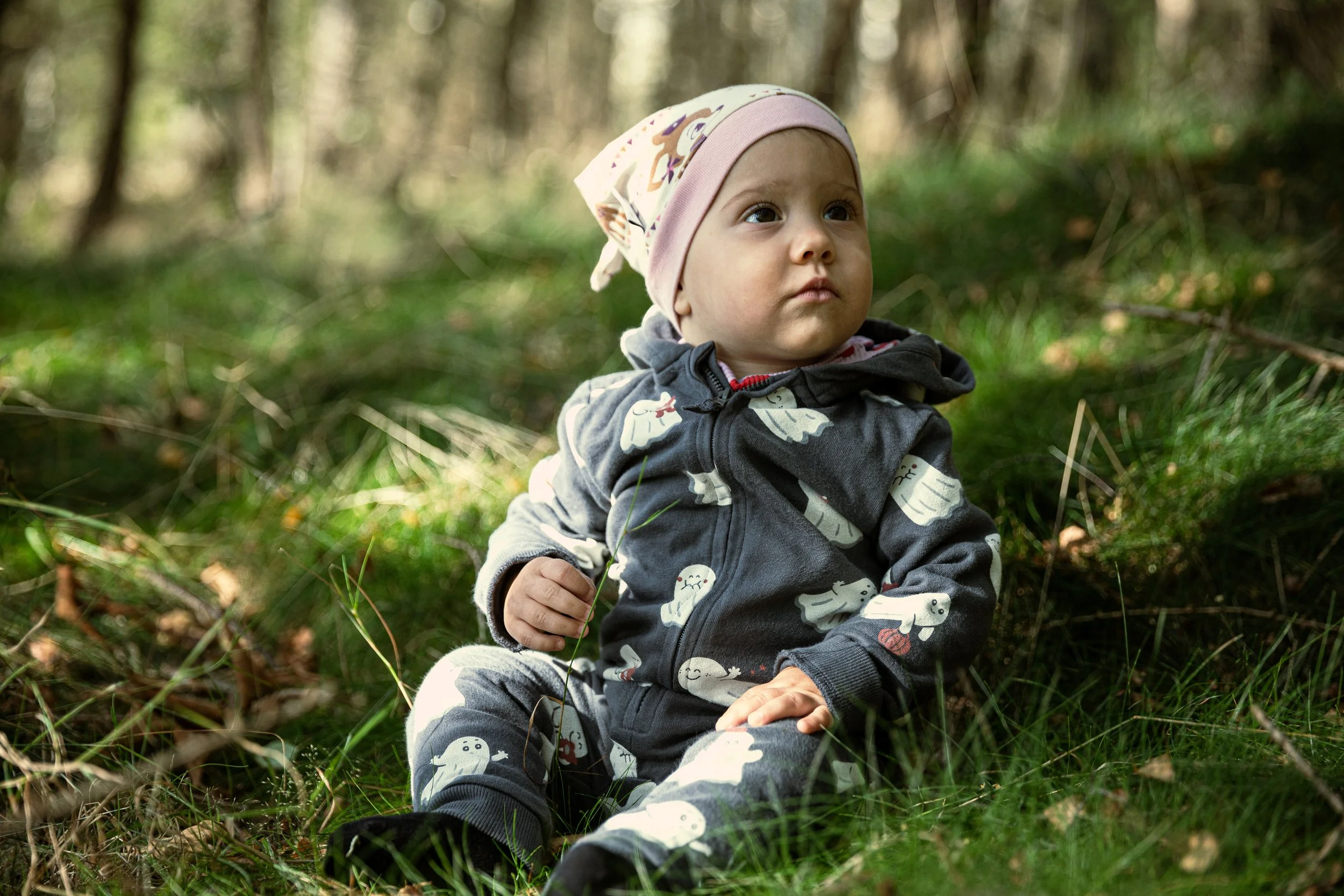 Ein kleiner Junge sitzt im Wald auf dem Boden, trägt eine Mütze und einen Hoodie mit Tiermuster, umgeben von grünen Zweigen und Bäumen.
