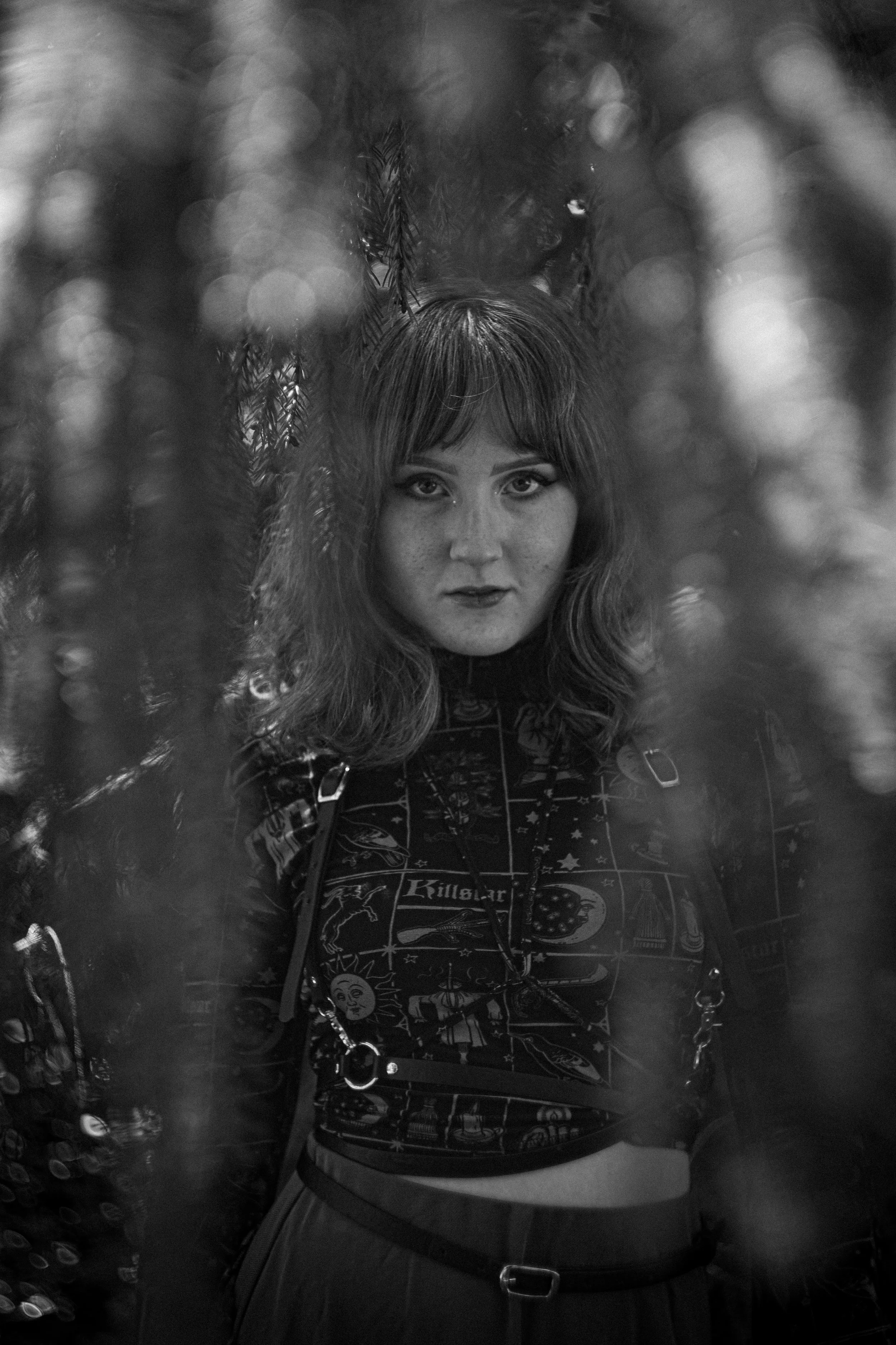 Schwarz-Weiß-Foto eines Mädchens mit langen Haaren, dunklem Make-up und einem kreativen Outfit, das Fotos am Hals trägt, umgeben von Bäumen und Blättern.