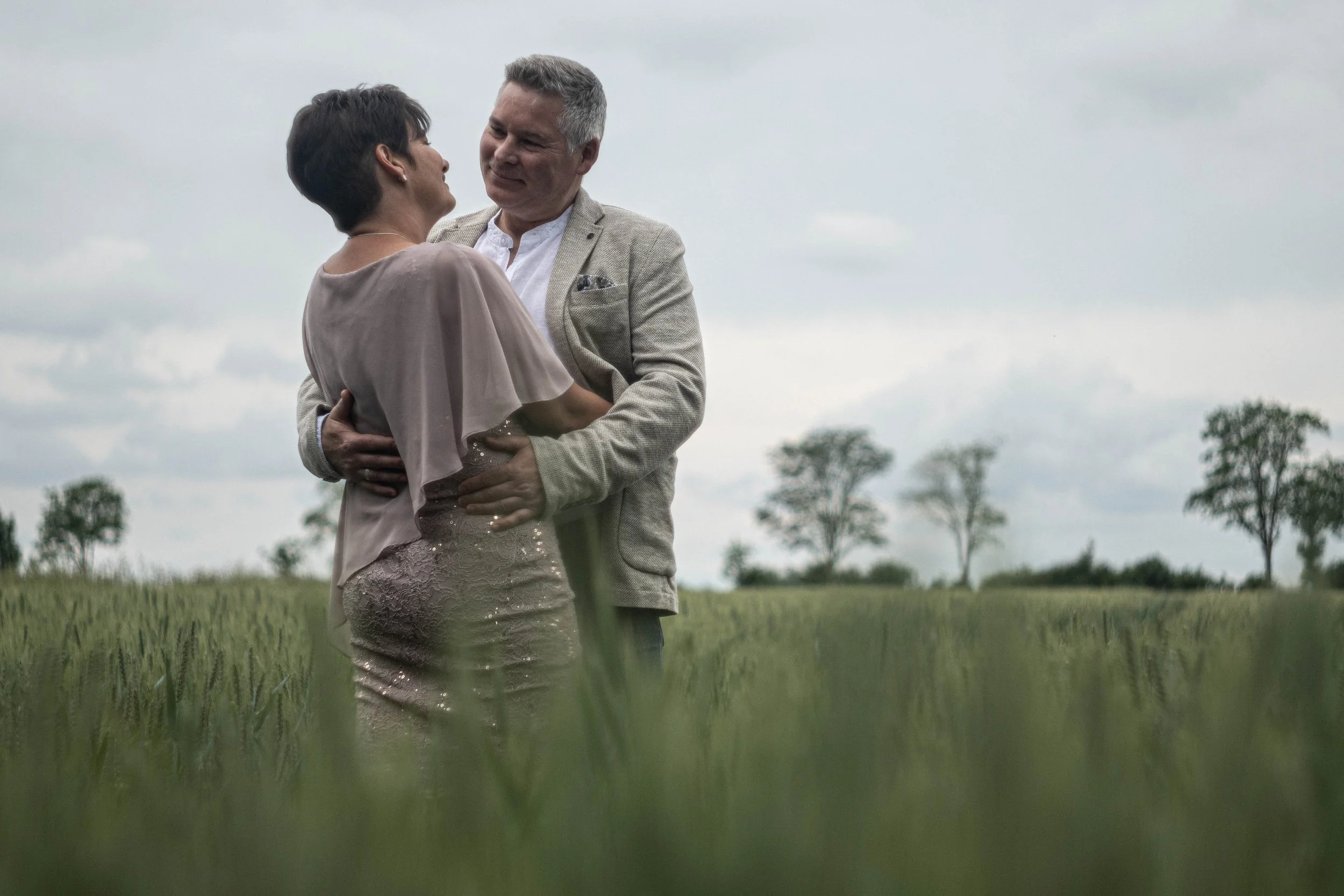 Ein Mann und eine Frau tanzen zusammen im Freien auf einem Feld, umgeben von Bäumen und unter einem bewölkten Himmel.