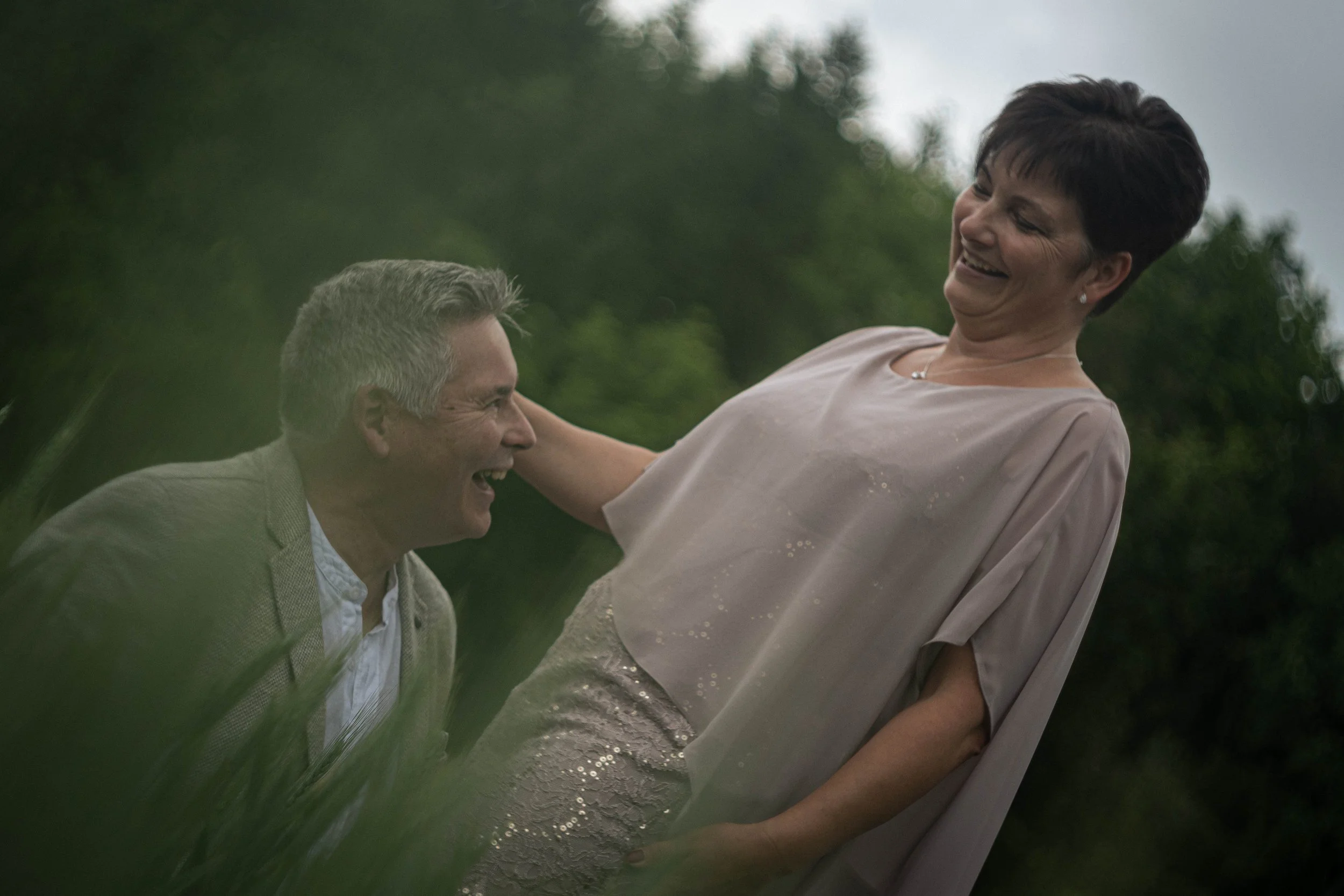 Ein Mann und eine Frau lachen zusammen im Wasser, während die Frau vom Mann gehalten wird, bei bewölktem Himmel im Freien.