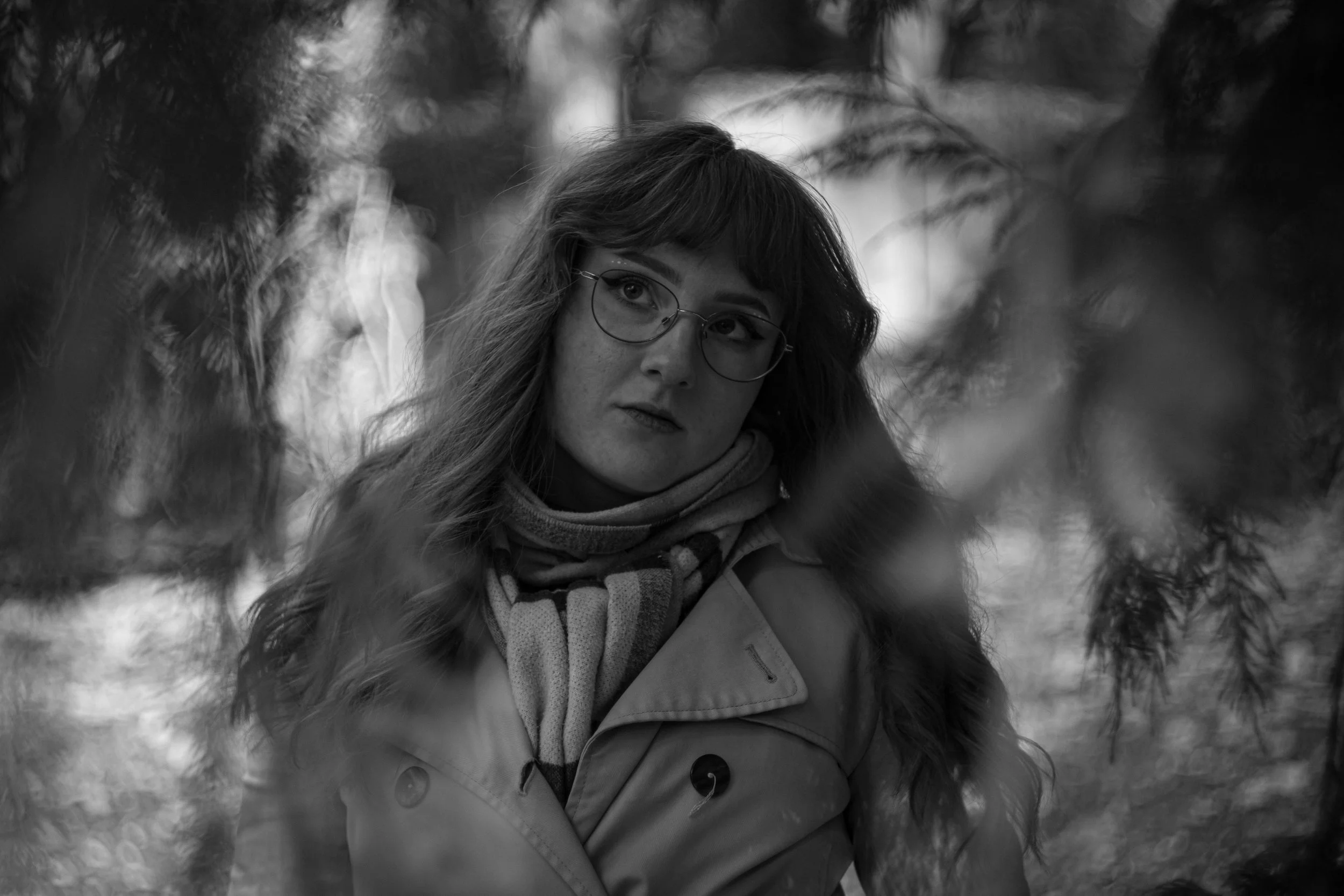 Eine junge Frau mit Brille und Mehrfarbiger Schal trägt eine Winterjacke mit Pelzbesatz auf einem Waldweg im Winter.