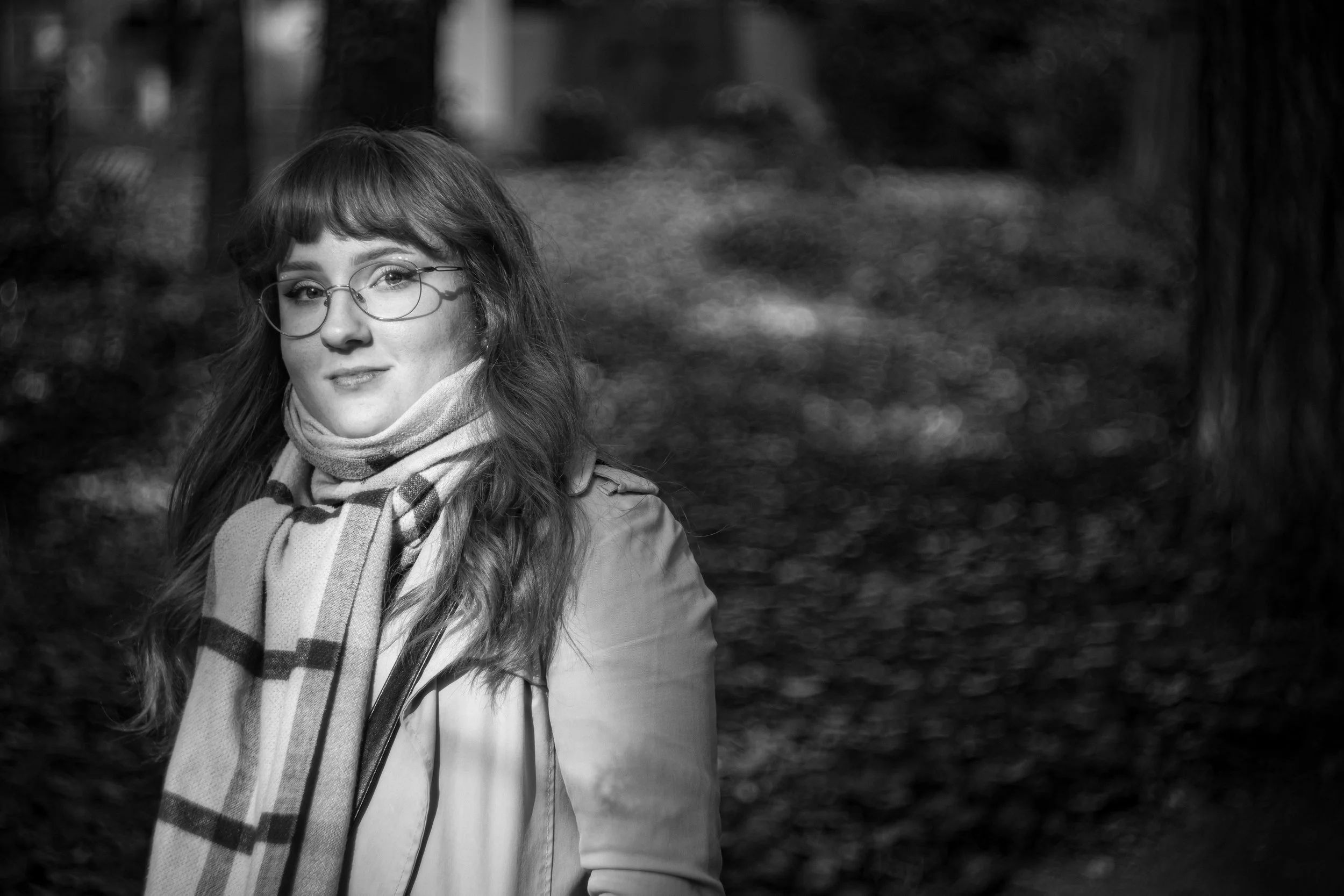 Schwarz-weiß-Foto eines jungen Mädchens mit Brille, langen Haaren, Schal und Jacke im Freien, im Hintergrund Bäume und Laub.