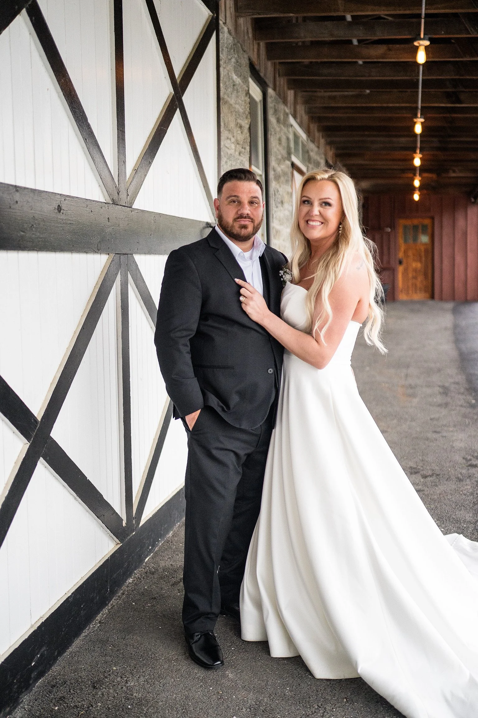A newlywed couple standing in front of a barn wall at night. The groom is wearing a black suit and white shirt, and the bride is in a white wedding gown with long blonde hair, both smiling.