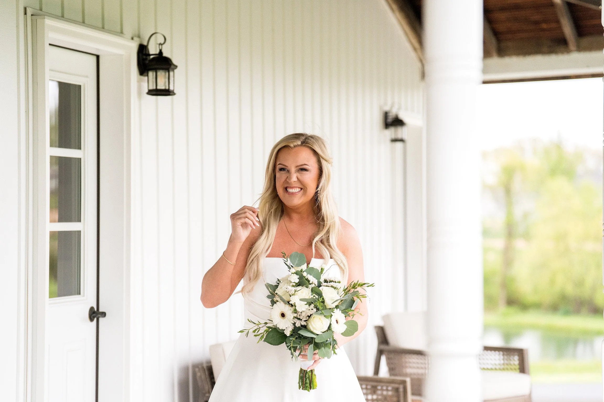 A smiling woman in a white dress holding a bouquet of white flowers and greenery, standing outside a porch with white walls and black outdoor light fixtures.