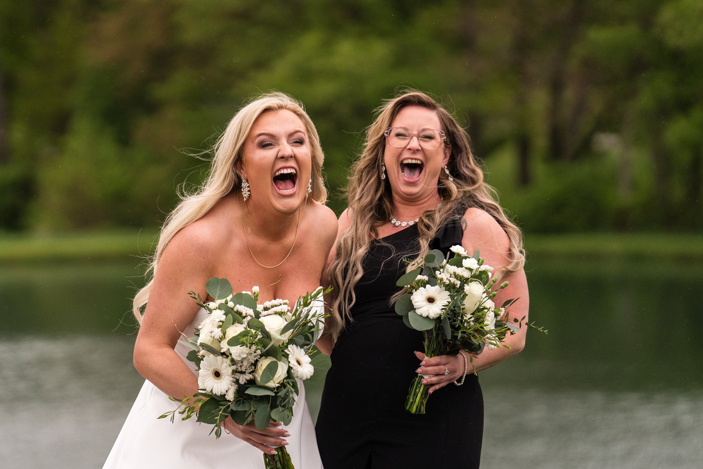 Two women, dressed for a wedding, are smiling and laughing outdoors while holding bouquets of white flowers and greenery, with a blurred green background.