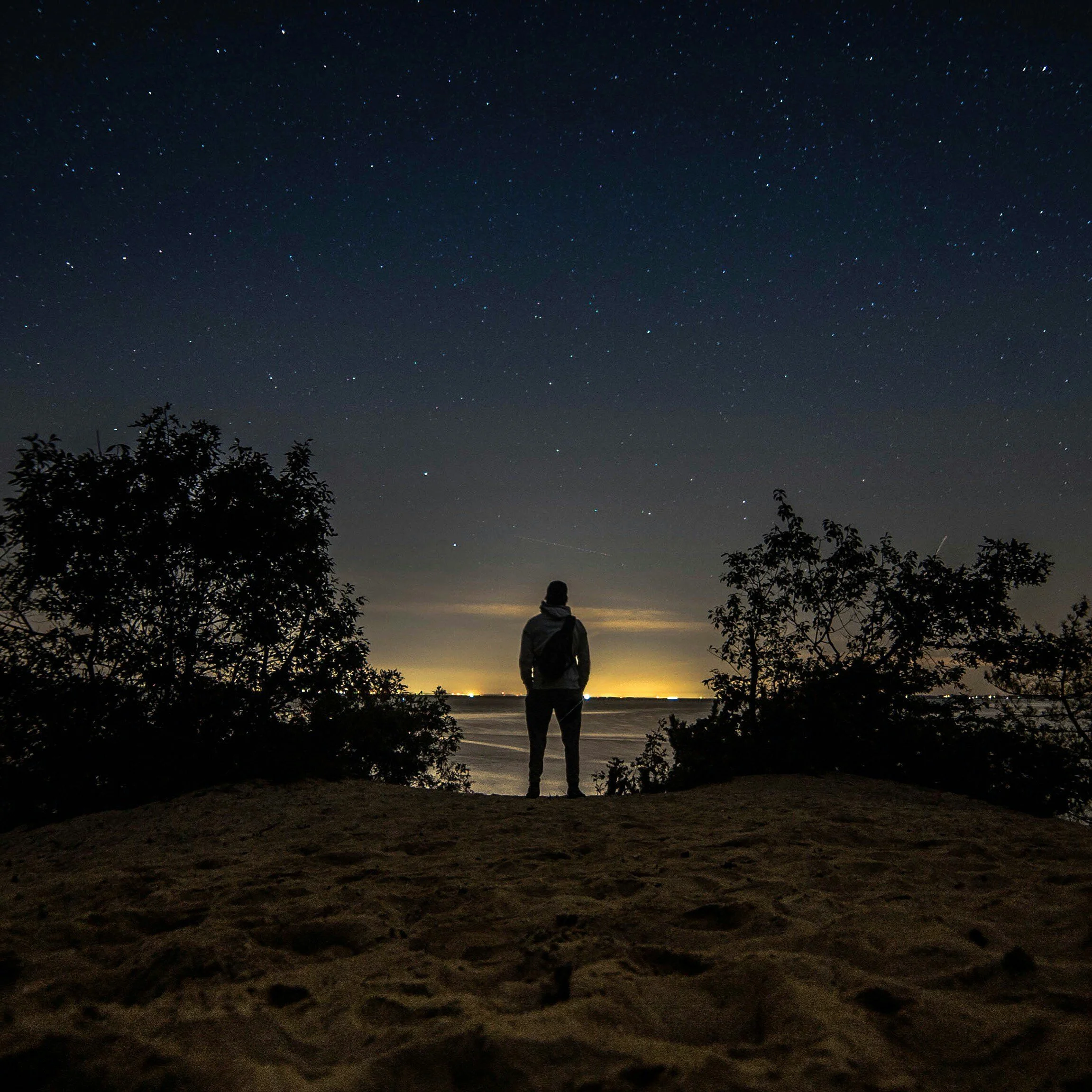 A silhouette of a person standing on a beach at night, looking at a star-filled sky over the ocean.