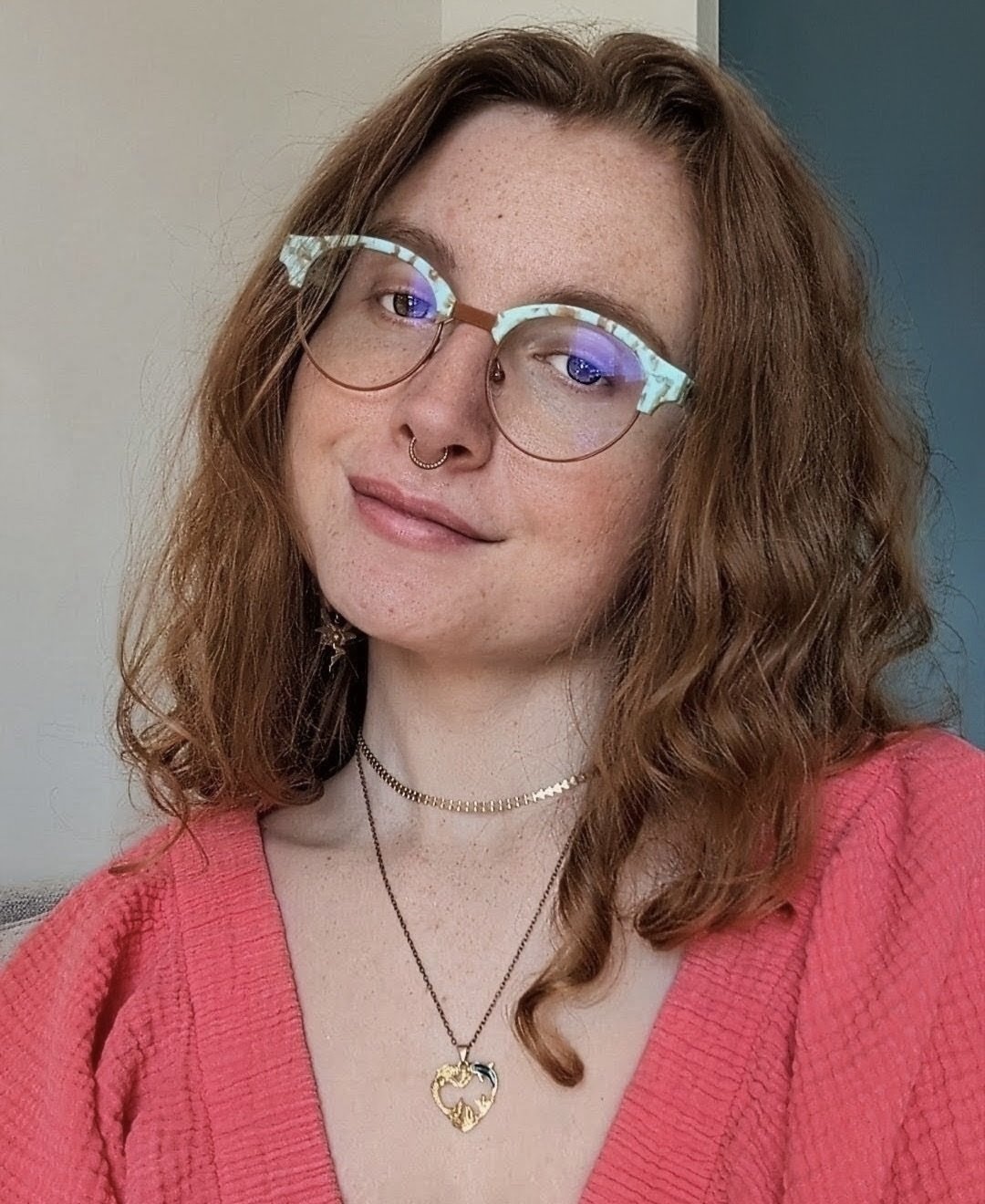 A young woman with curly red hair, wearing glasses with light green frames, a septum piercing, layered necklaces, and a pink sweater, smiling at the camera indoors.