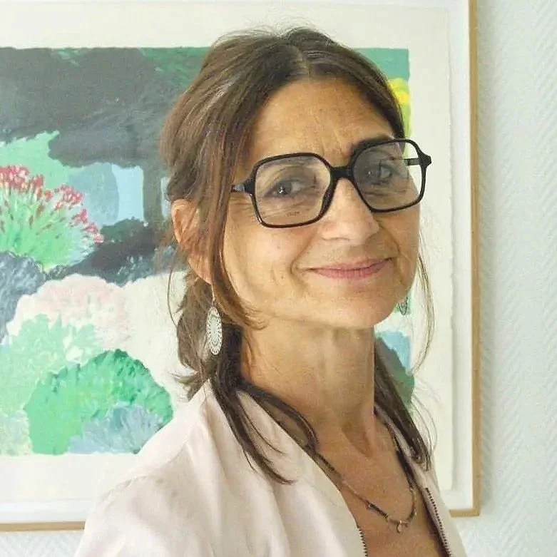 Une femme souriante portant des lunettes et des boucles d'oreilles, avec un tableau abstrait en arrière-plan.