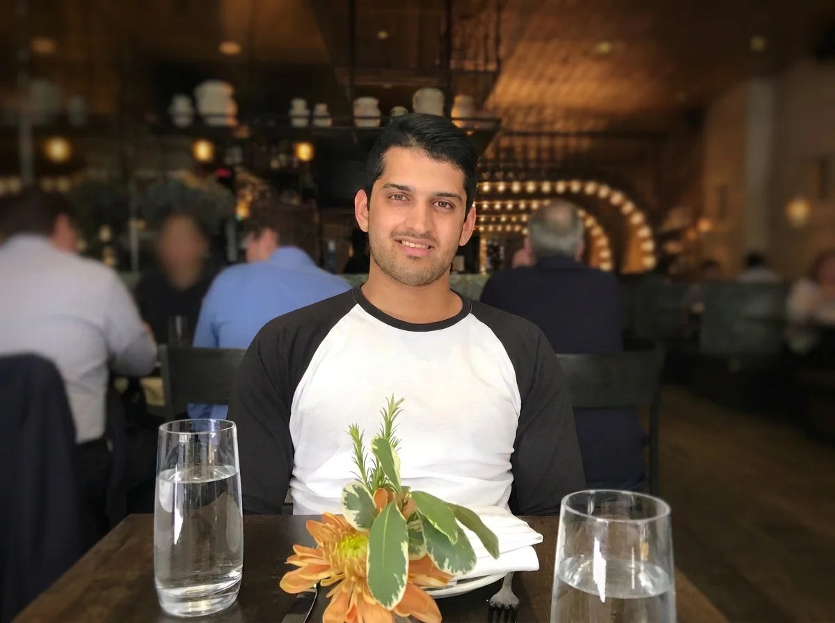 A young man sitting at a restaurant table with two glasses of water, a plate, a napkin, and a floral centerpiece in front of him, smiling at the camera.