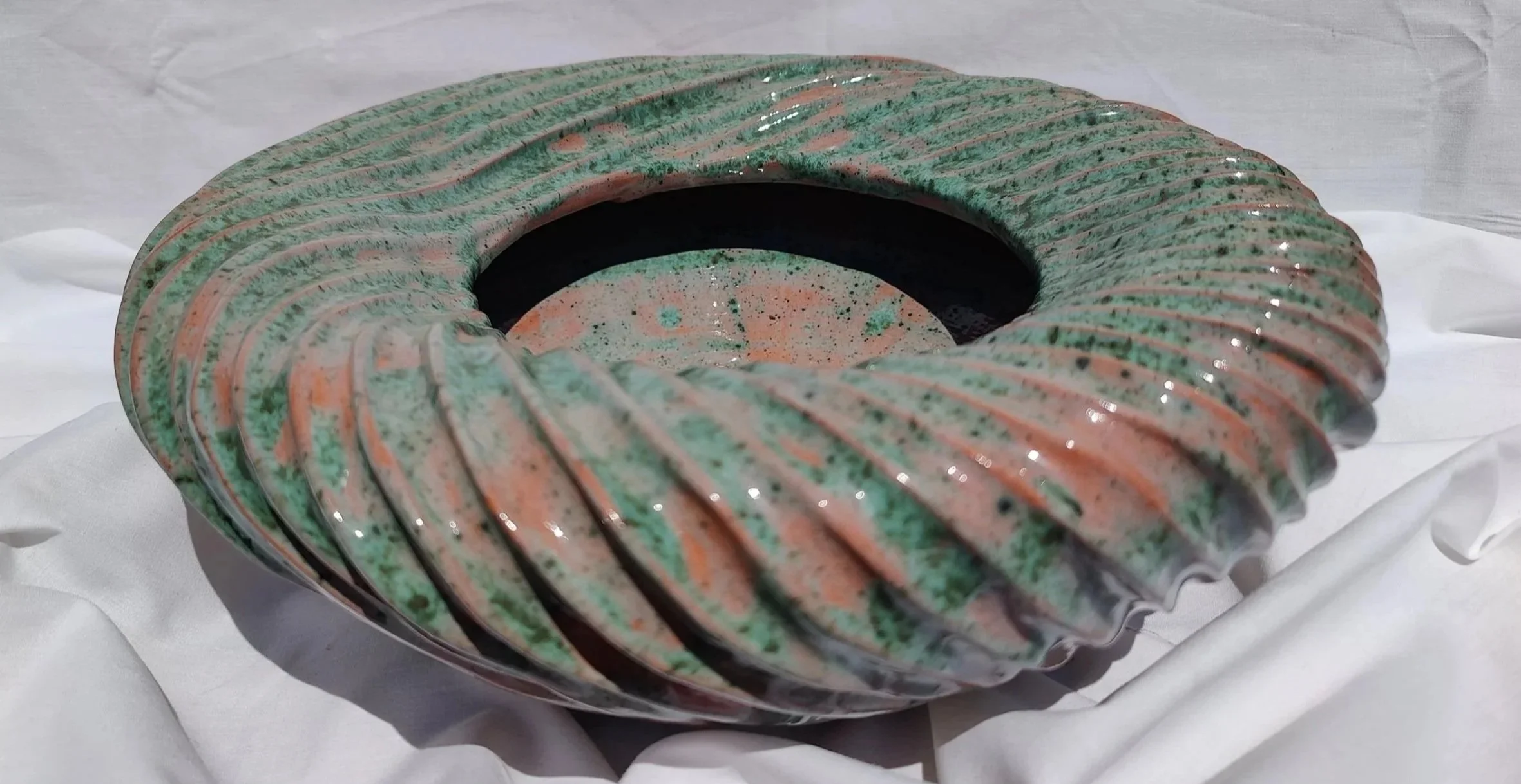 Ceràmica decorativa en forma toroidal amb textura acanalada i colors en tons verds, rosats i negres, col·locada sobre una superficie blanca i opaca.