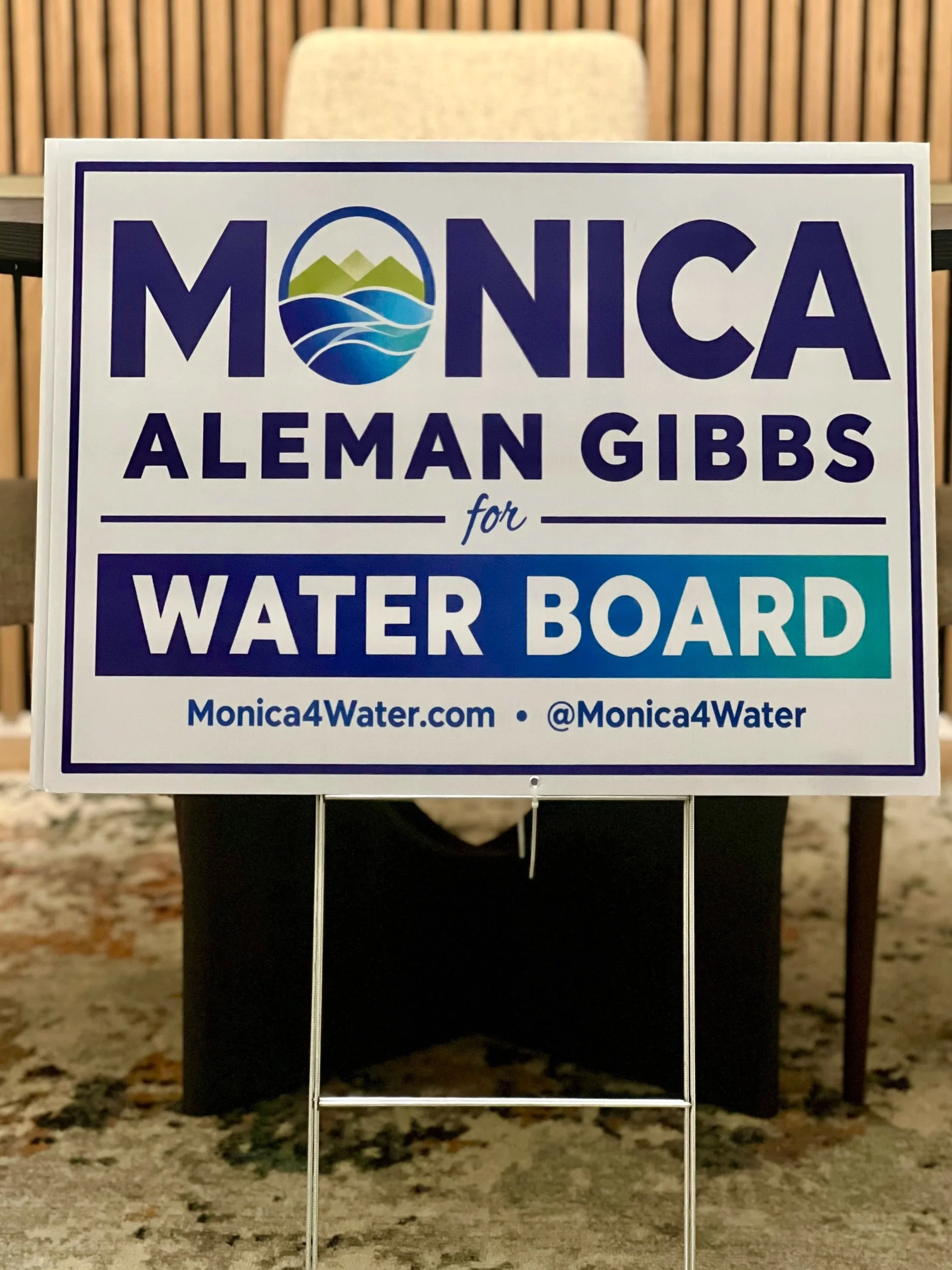 A sign with the name Monica Alemán Gibbs for Water Board placed on a stand. The background includes a wooden wall and a chair.
