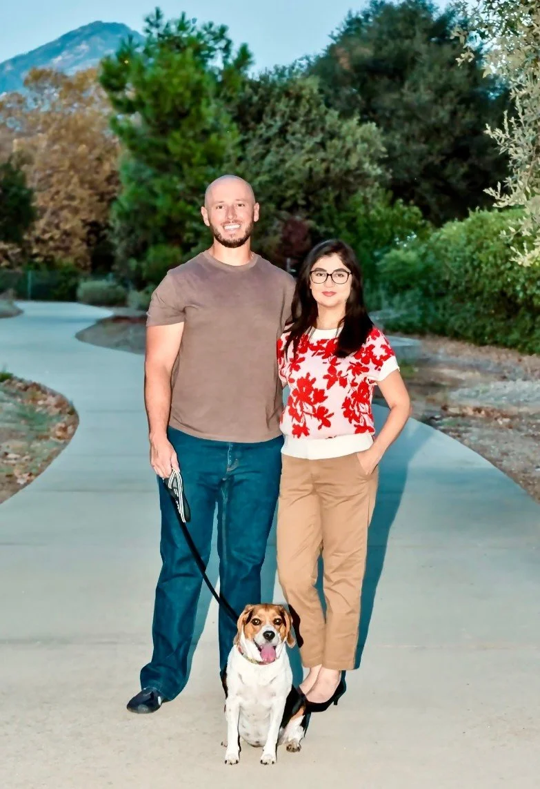 A smiling man and woman standing on a sidewalk outdoors with a small dog sitting in front of them. The man has a beard, is wearing a brown t-shirt and jeans, and holds the dog’s leash. The woman has dark hair, glasses, and is wearing a floral red and white top with beige pants. The background features lush green trees and a mountain in the distance.