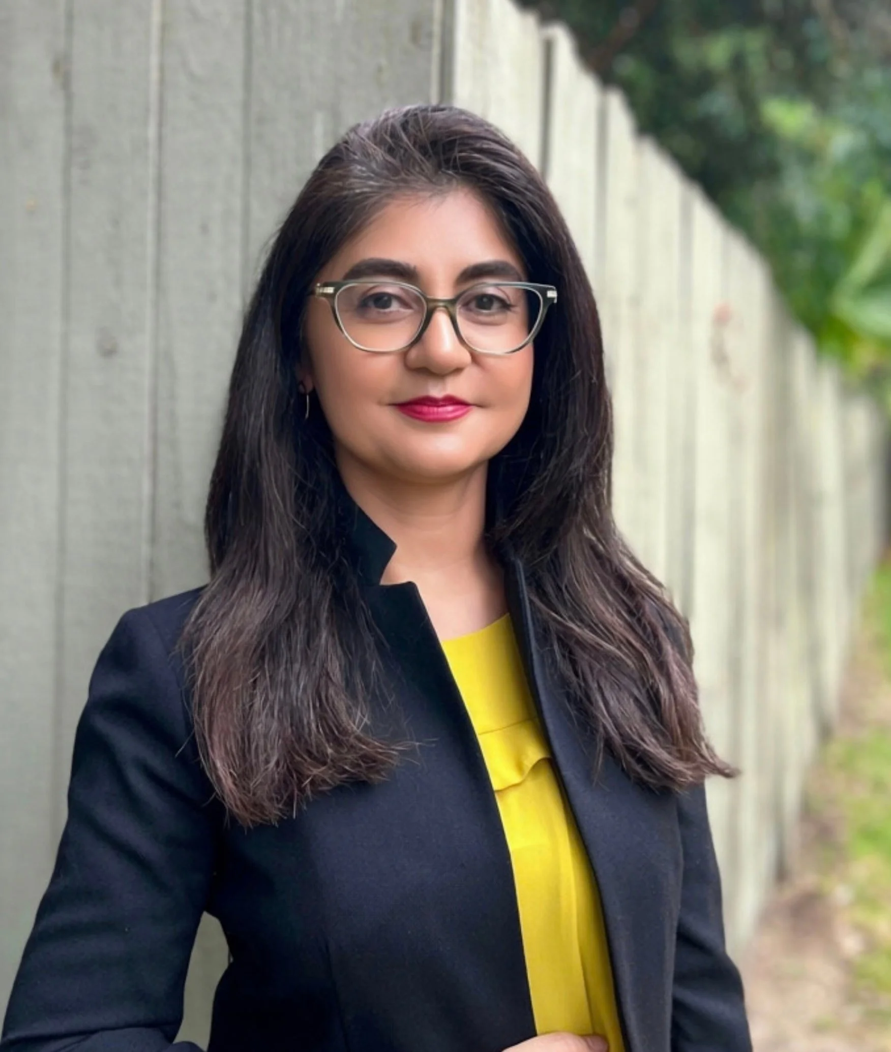 Monica aleman Gibbs. water board candidate in black blazer and yellow blouse wearing glasses standing against a fence.