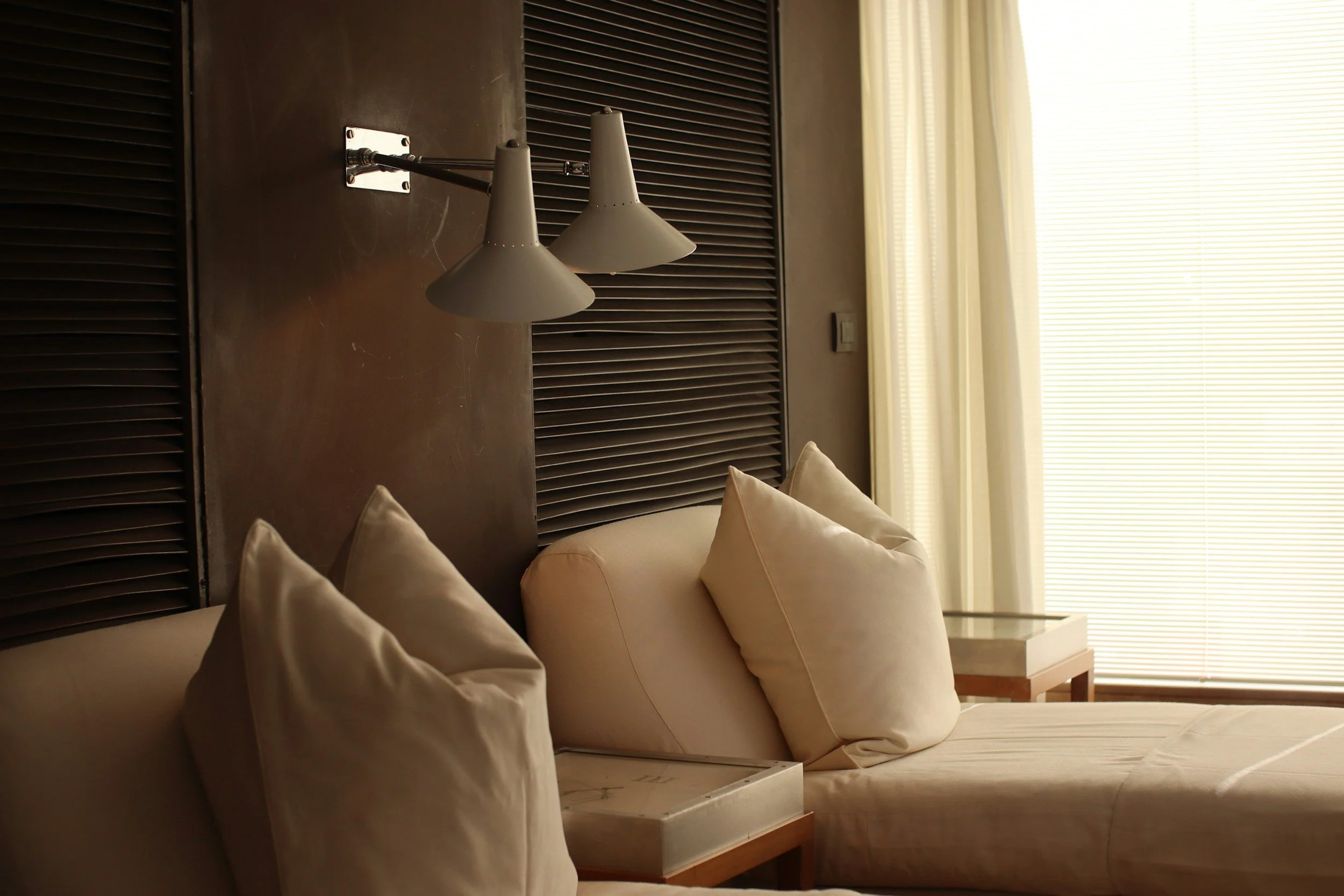 A bedroom corner with beige pillows on a bed, a white nightstand, an adjustable wall lamp, dark wooden cabinet doors, and a window with closed horizontal blinds letting in sunlight.