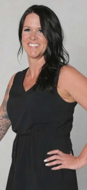Woman with black hair, smiling, wearing a black sleeveless jumpsuit, with tattoos on her left arm, standing against a plain gray background.