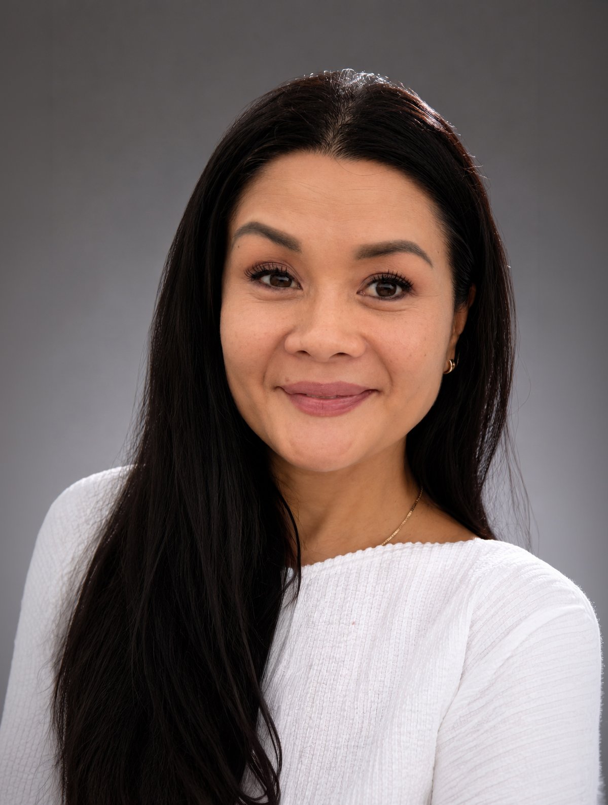 A woman with long black hair wearing a white top and gold earrings, smiling against a gray background.