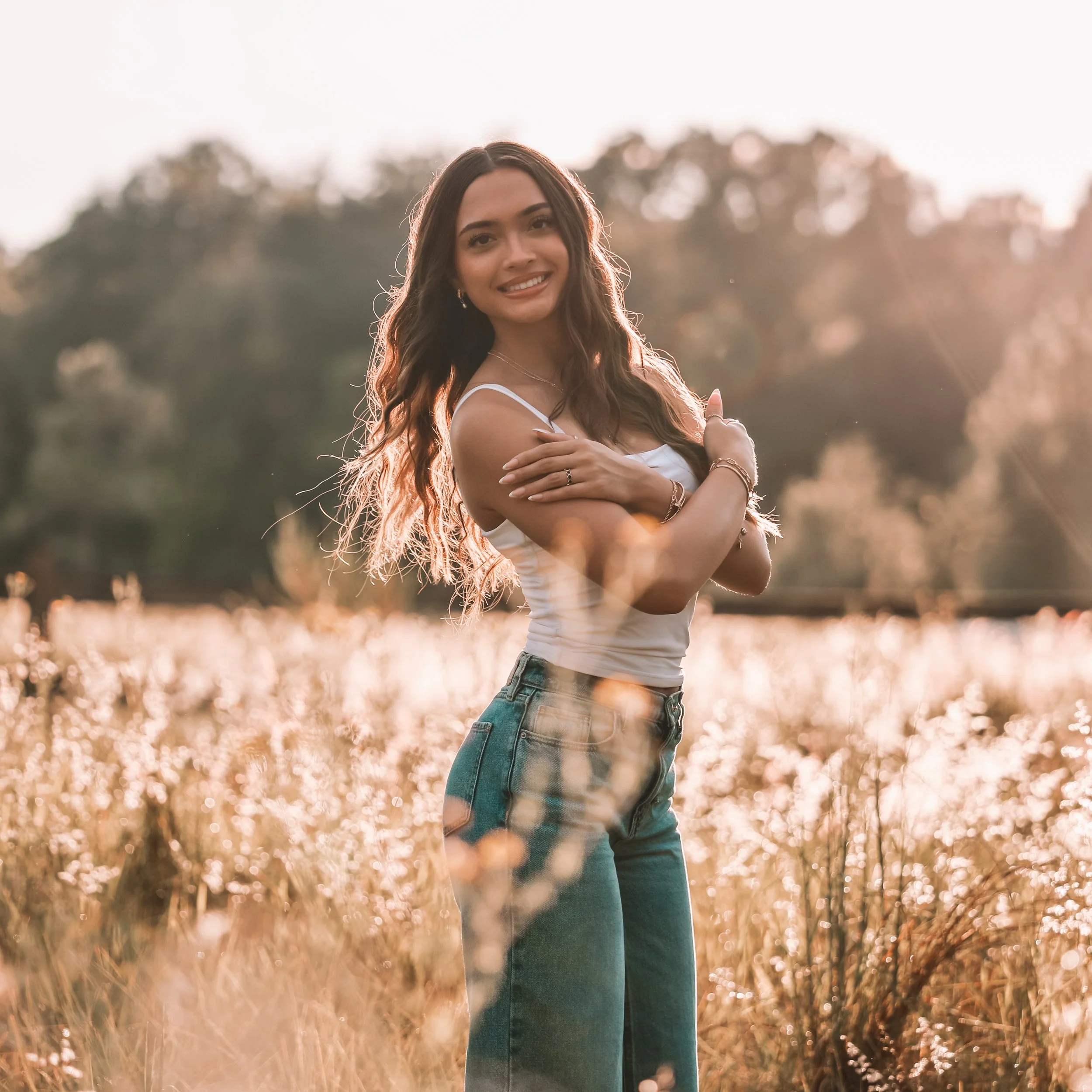 A young woman with long, wavy hair standing in a field at sunset, smiling and crossing her arms over her chest.
