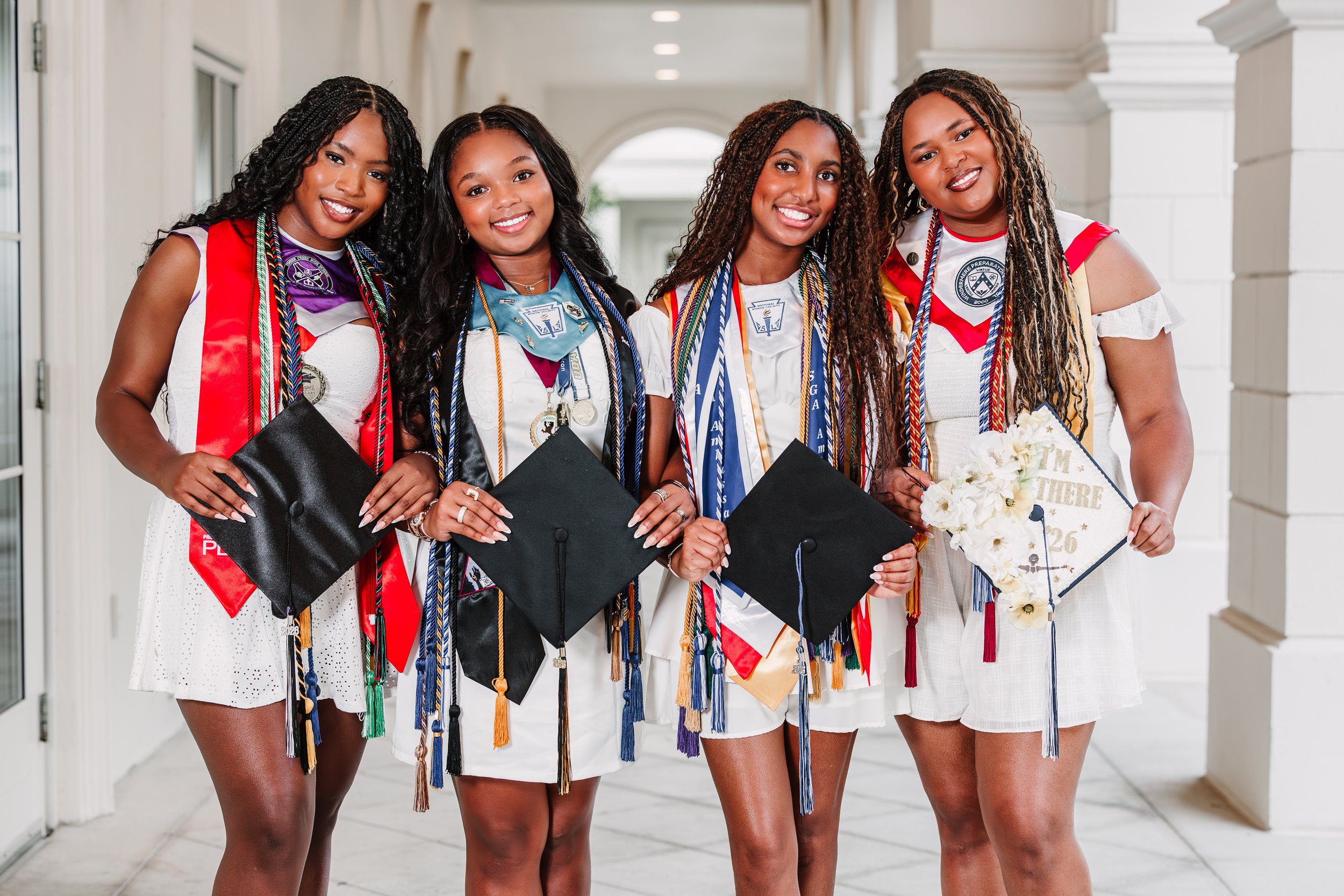 Four young women celebrating graduation, wearing caps, gowns, and academic honors, standing together in a hallway with smiling faces.