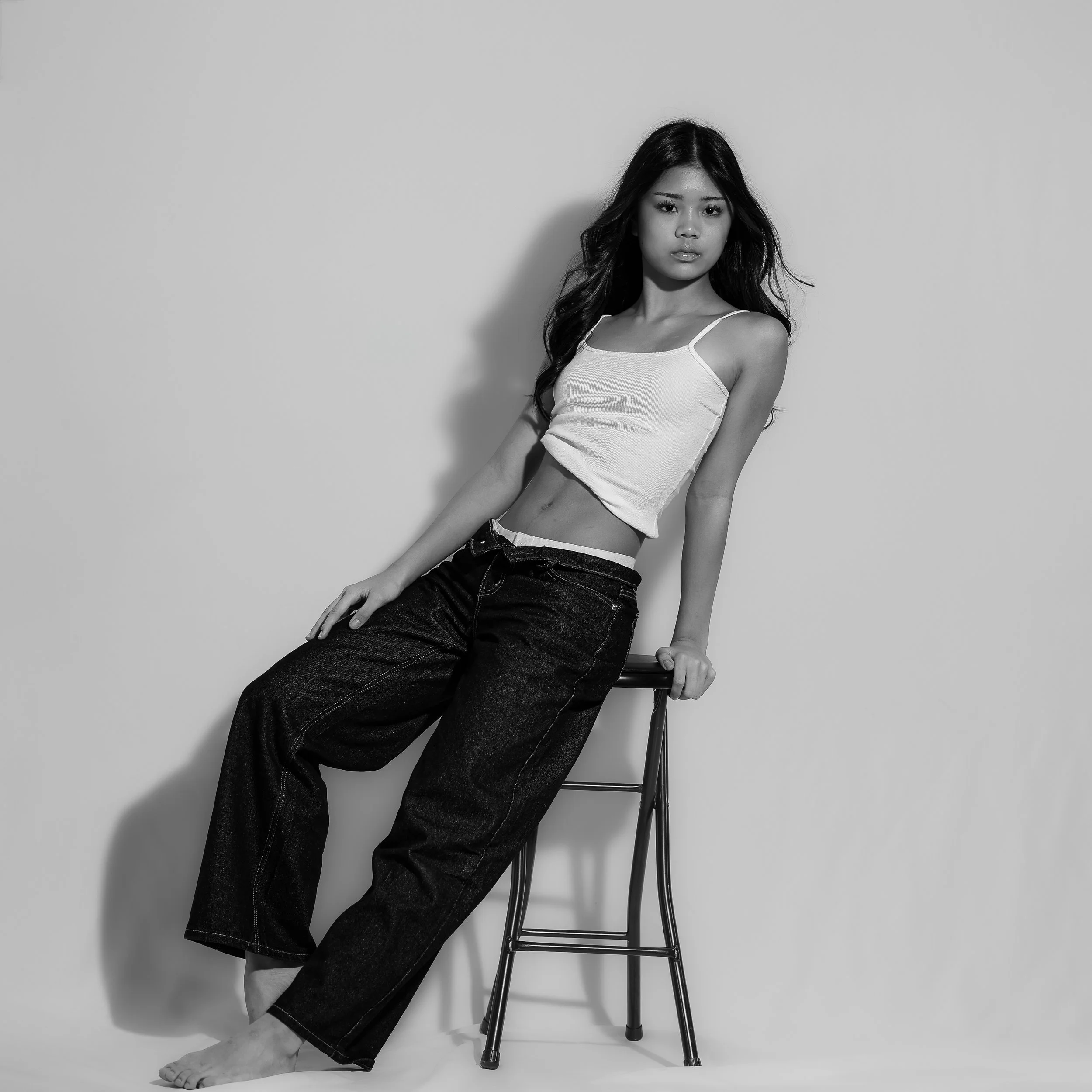 Black-and-white photo of a young woman with long wavy hair, wearing a tank top and jeans, sitting on a chair against a plain wall, with her right arm resting on her knee and her left arm hanging by her side.