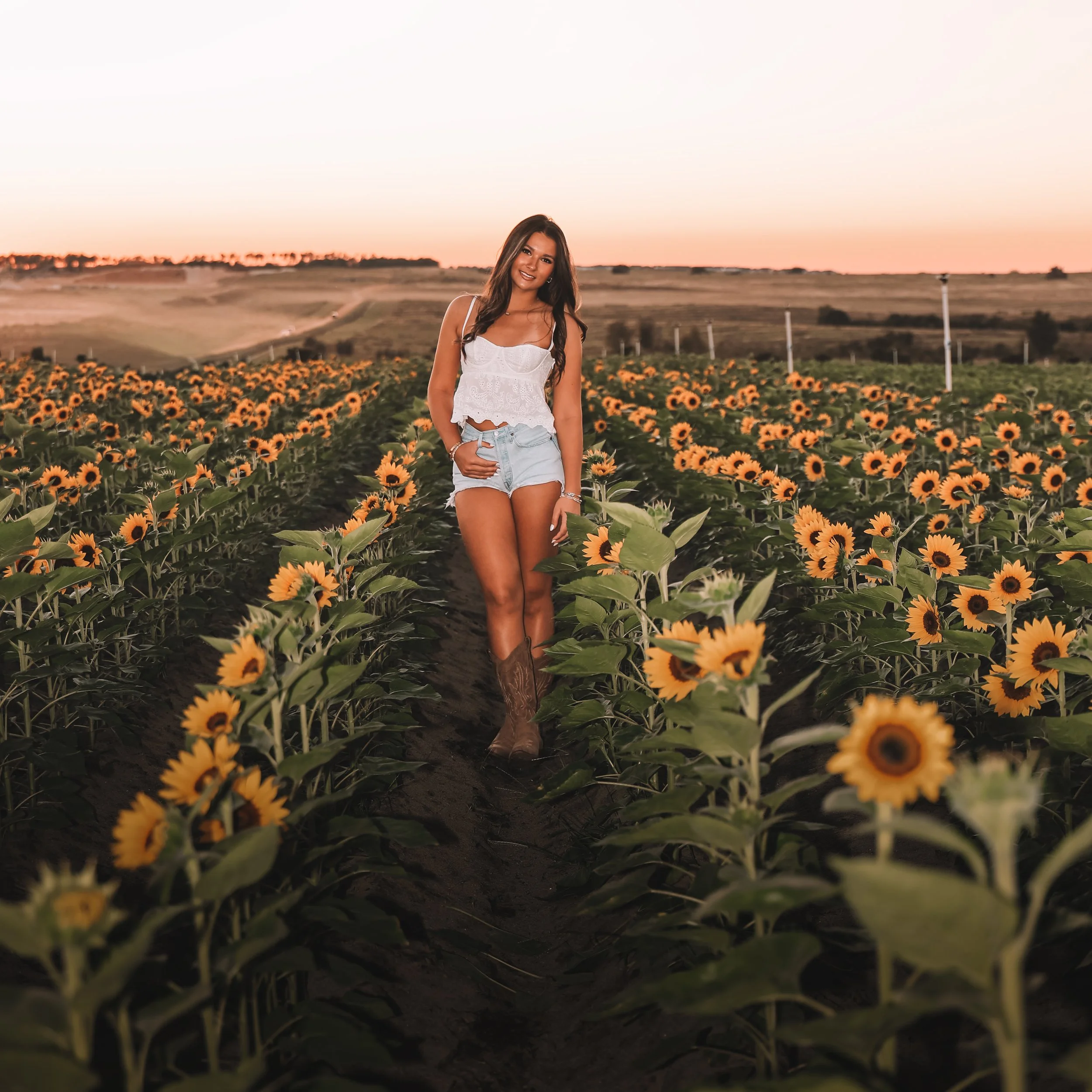 A young woman standing in a sunflower field at sunset, smiling and wearing a white crop top, denim shorts, and cowboy boots.