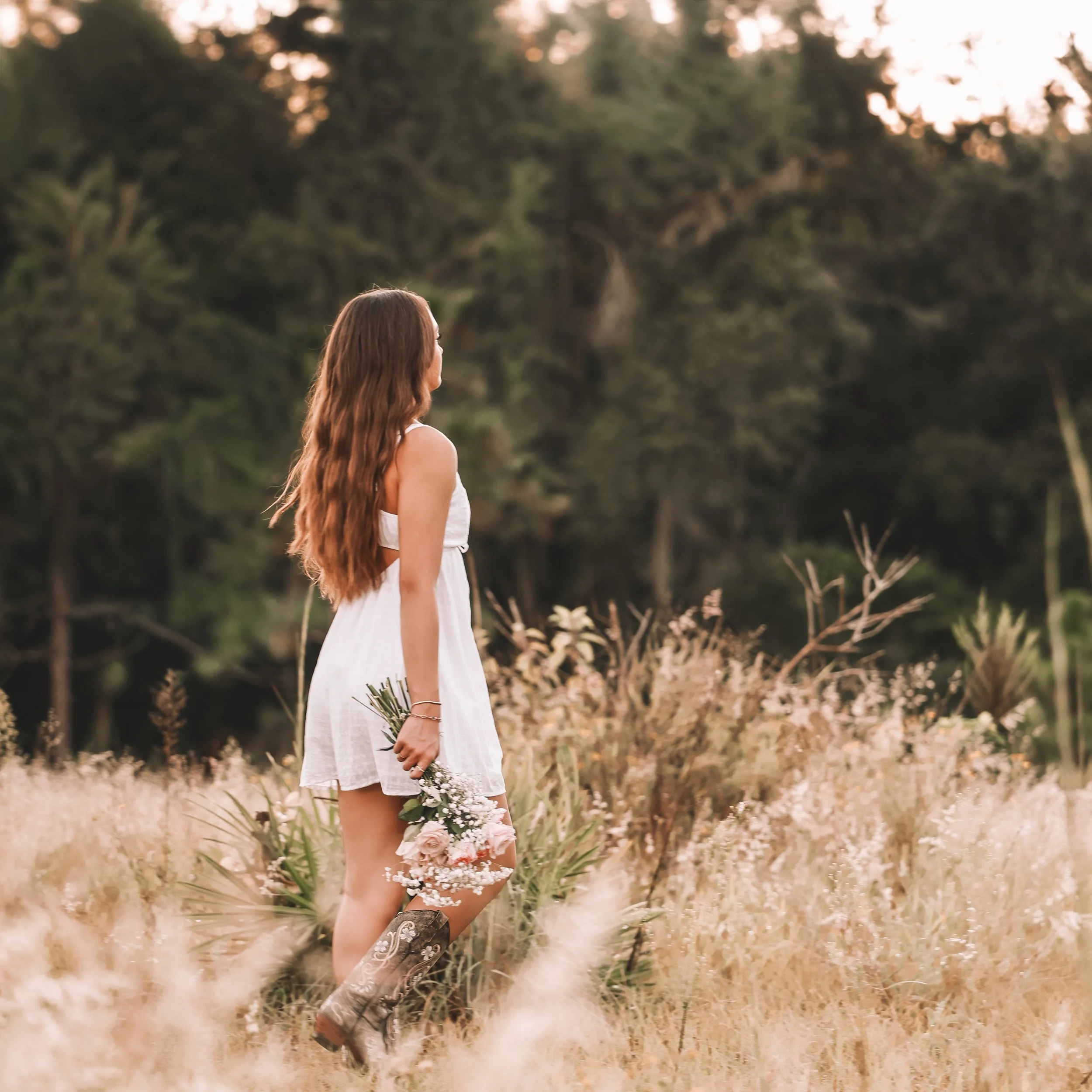 A woman with long, wavy hair wearing a white dress and cowboy boots standing in a field of tall grass and wildflowers, holding a small bouquet of flowers, with trees and a sunset sky in the background.