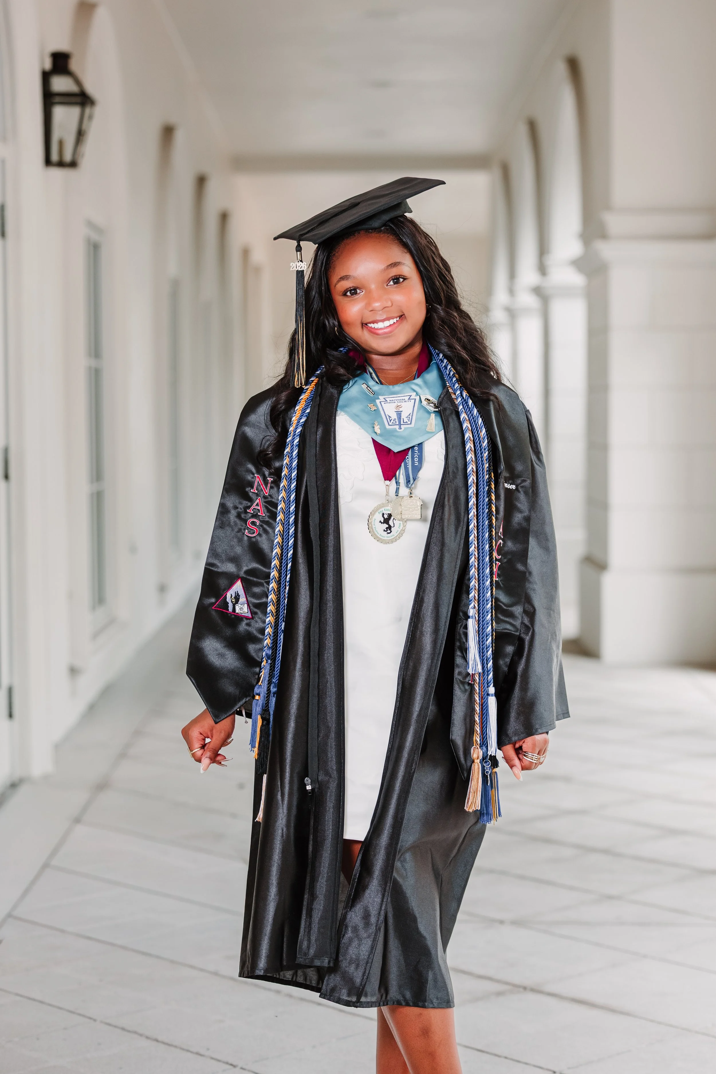 Young woman in graduation gown and cap standing in a bright hallway, smiling and looking at the camera, with medals and cords around her neck.