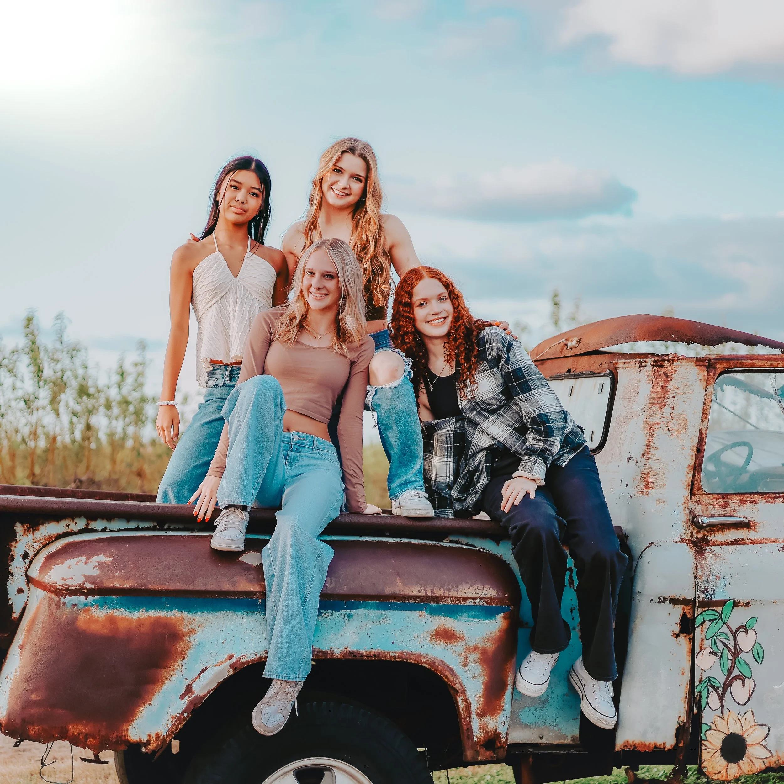 Group of five young women sitting and standing on a rusty, old food truck decorated with flowers and colorful paint outdoors in a field with blue sky and clouds.