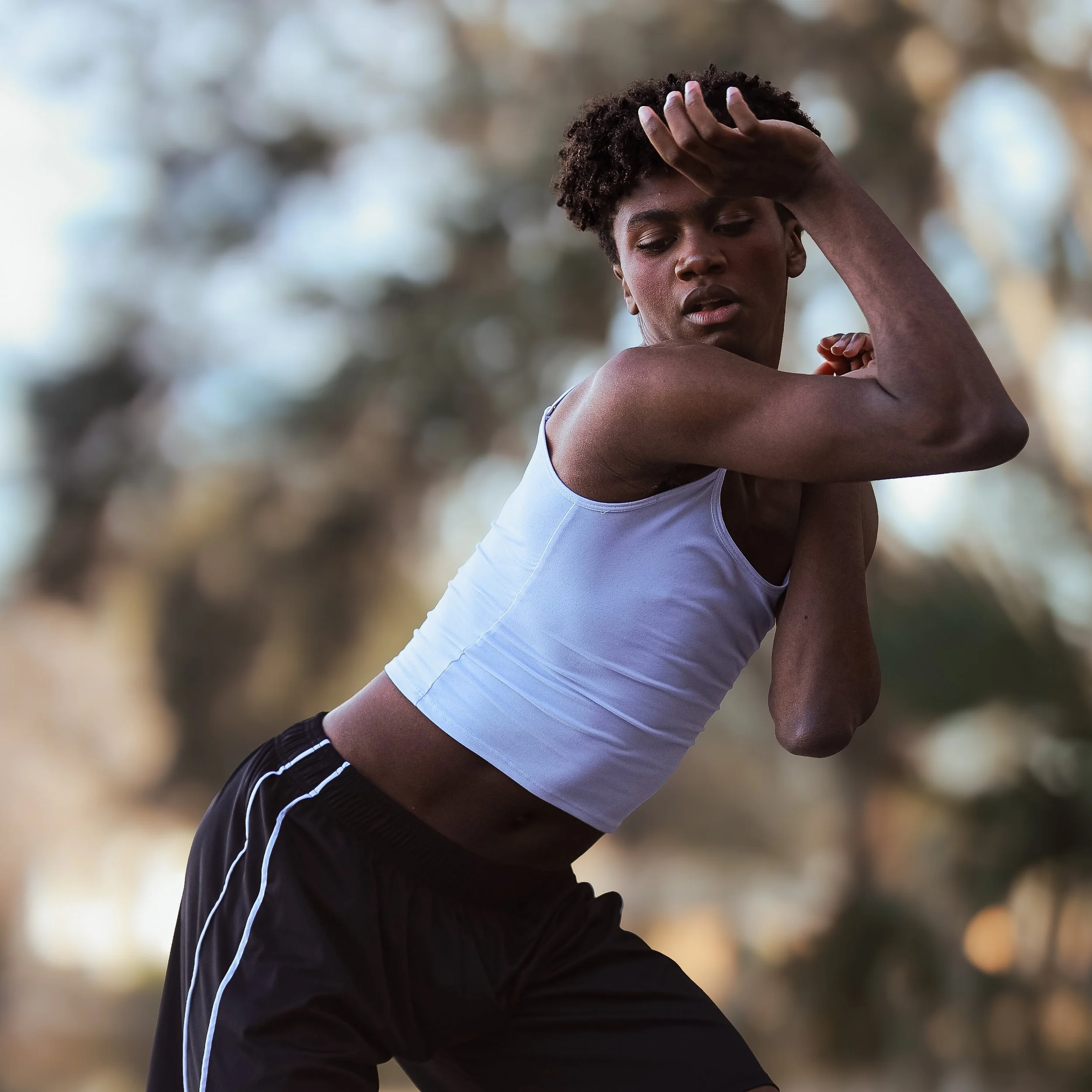 Young man with curly hair stretching outdoors during daytime, wearing a white tank top and black athletic shorts.