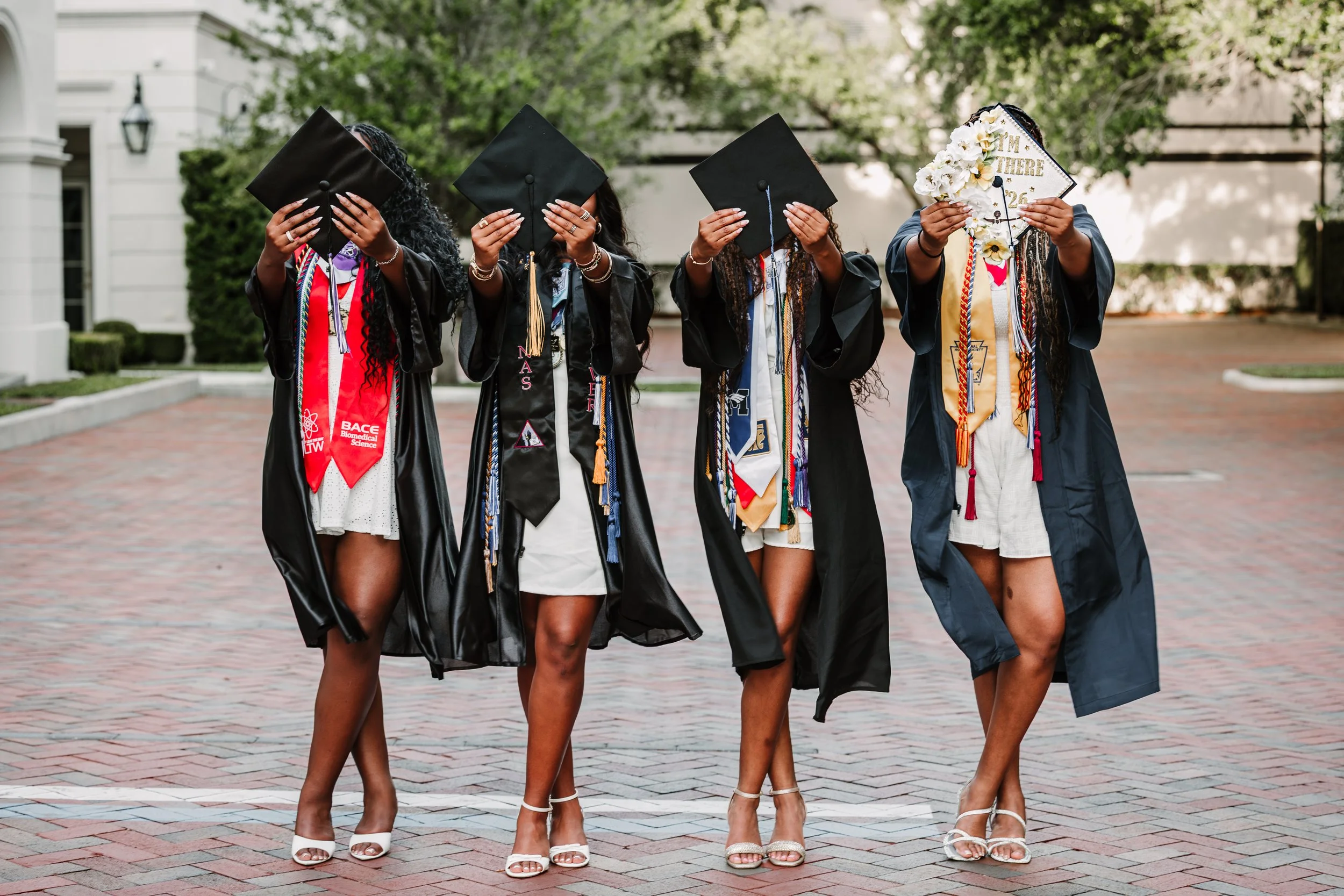 Group of five women in graduation robes and caps standing on a brick pathway with trees in the background, holding their caps covering their faces, with only their legs and shoes visible.