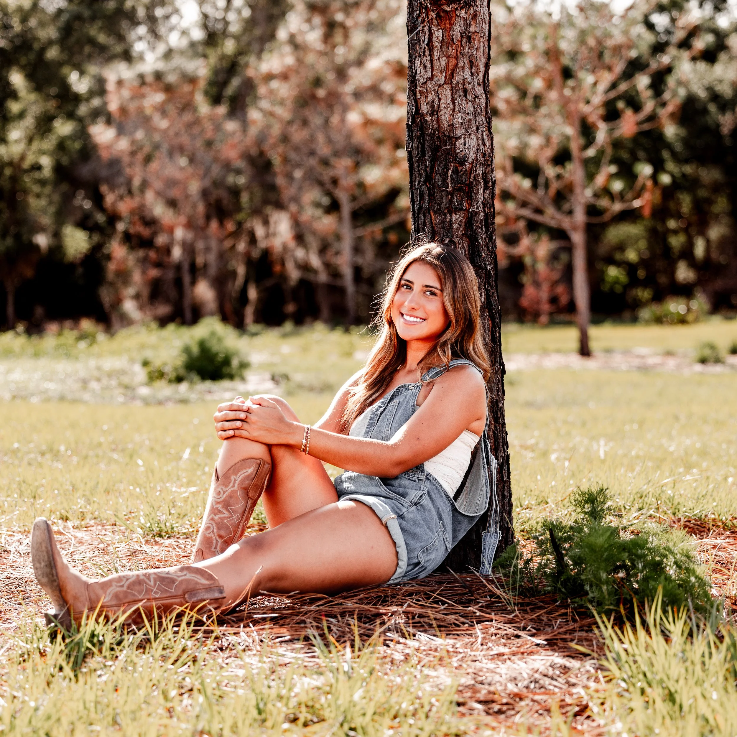 A young woman with long brown hair, sitting on the grass leaning against a tree, smiling, in a park with trees and grass in the background.