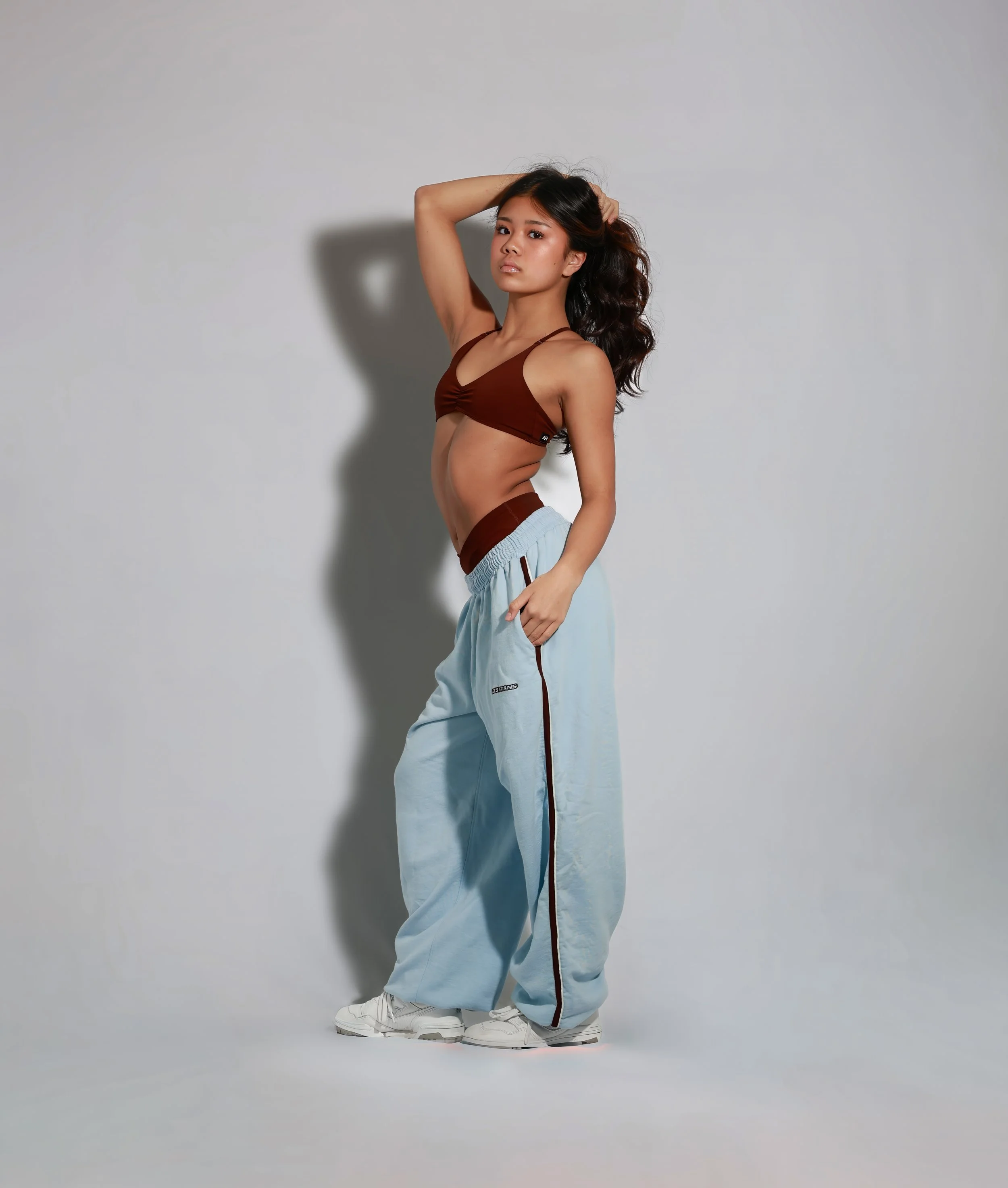Young woman with long wavy dark hair wearing a brown crop top, light blue baggy pants with red side stripes, white sneakers, standing against a gray background, with her hand on her head.