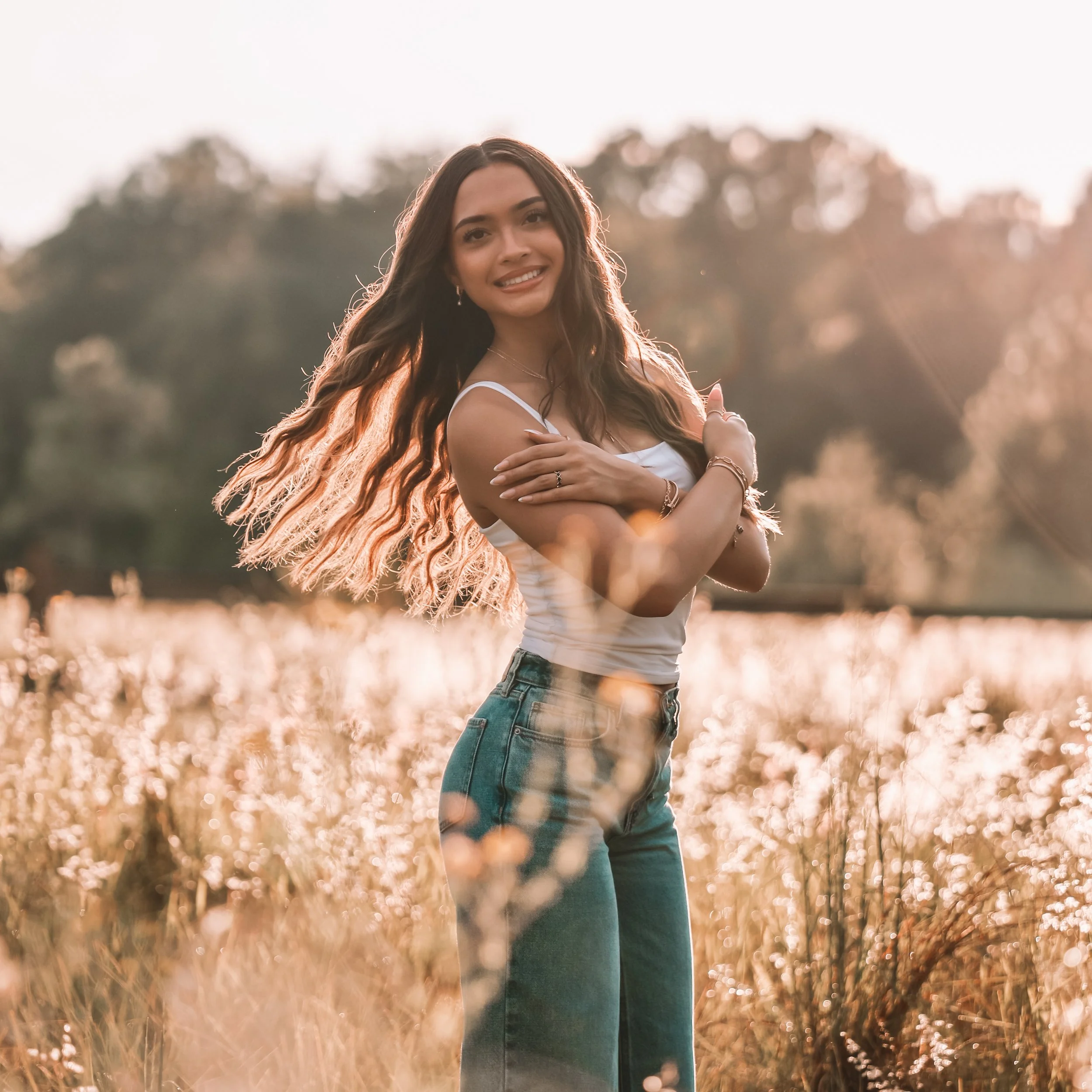 A smiling young woman with long, wavy brown hair, wearing a white tank top and jeans, standing in a field of tall grass during sunset.