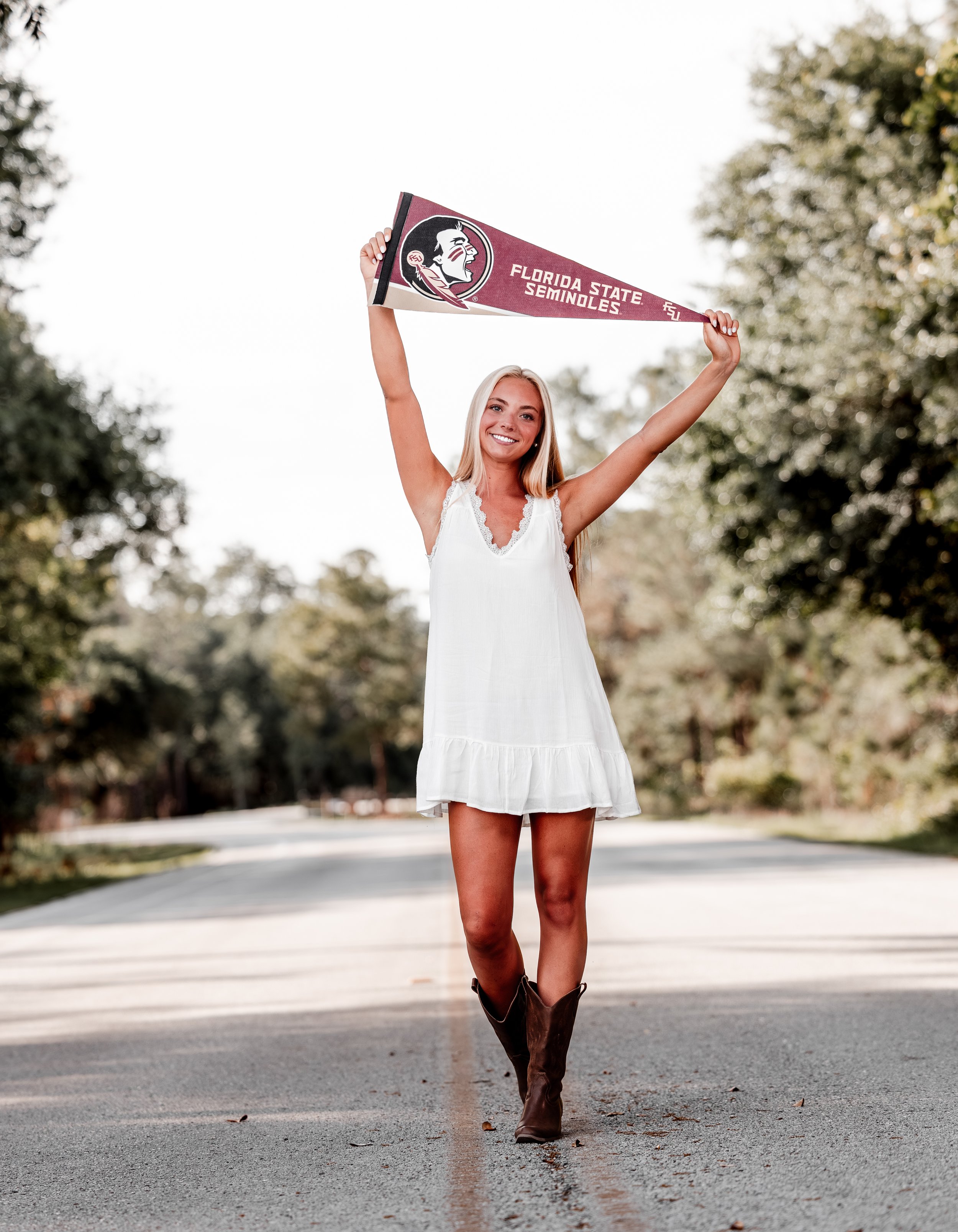 A woman in a white dress and brown cowboy boots holds a maroon Florida State Seminoles pennant above her head while walking on a paved path surrounded by trees. Senior Dance Photography - Orlando FL