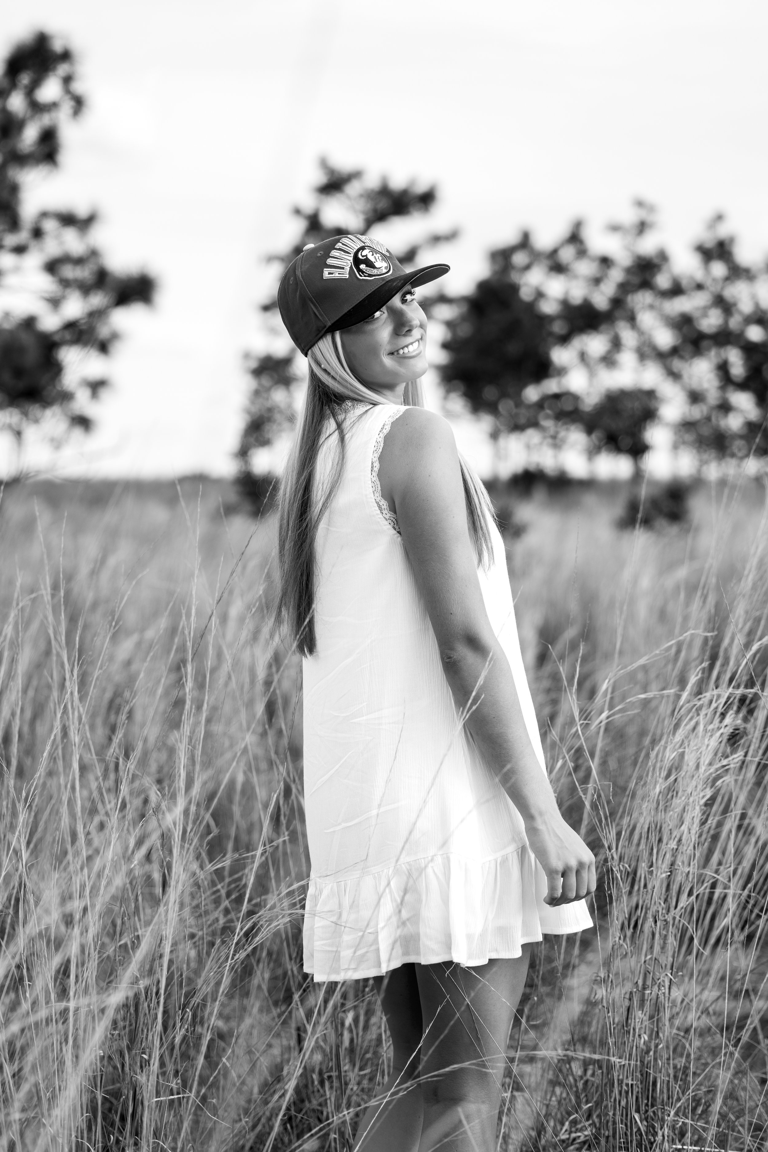 A smiling woman wearing a baseball cap and white dress standing in a field of tall grass with trees in the background.