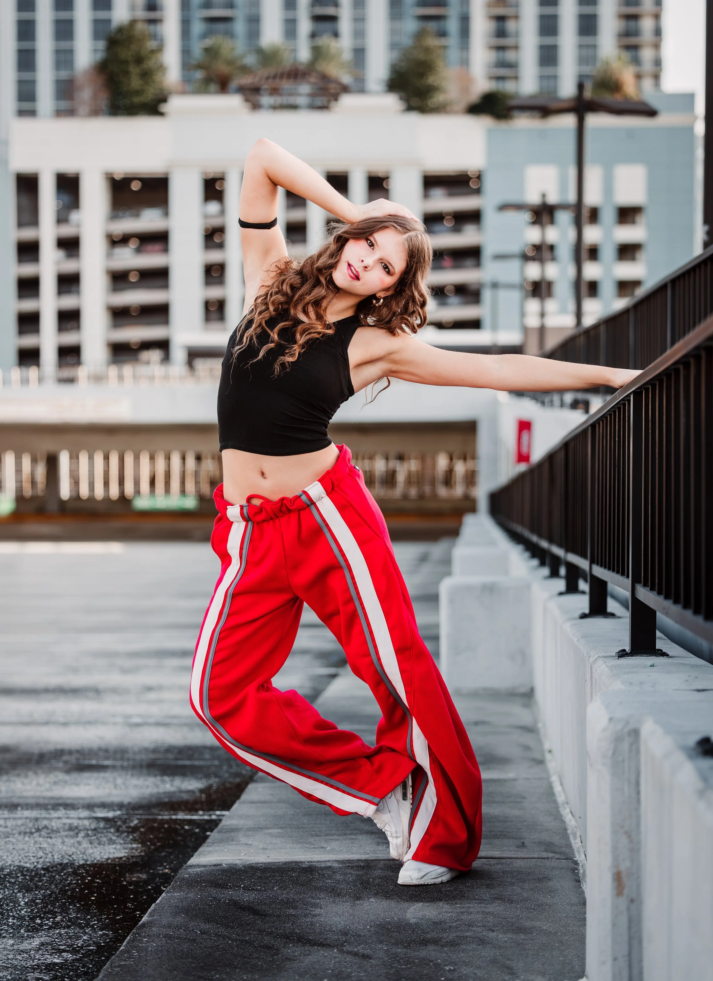 A woman with long, curly brown hair and red lipstick poses outdoors on a parking garage ledge, wearing a black crop top, red track pants with white and grey stripes, and white sneakers, with urban buildings in the background.