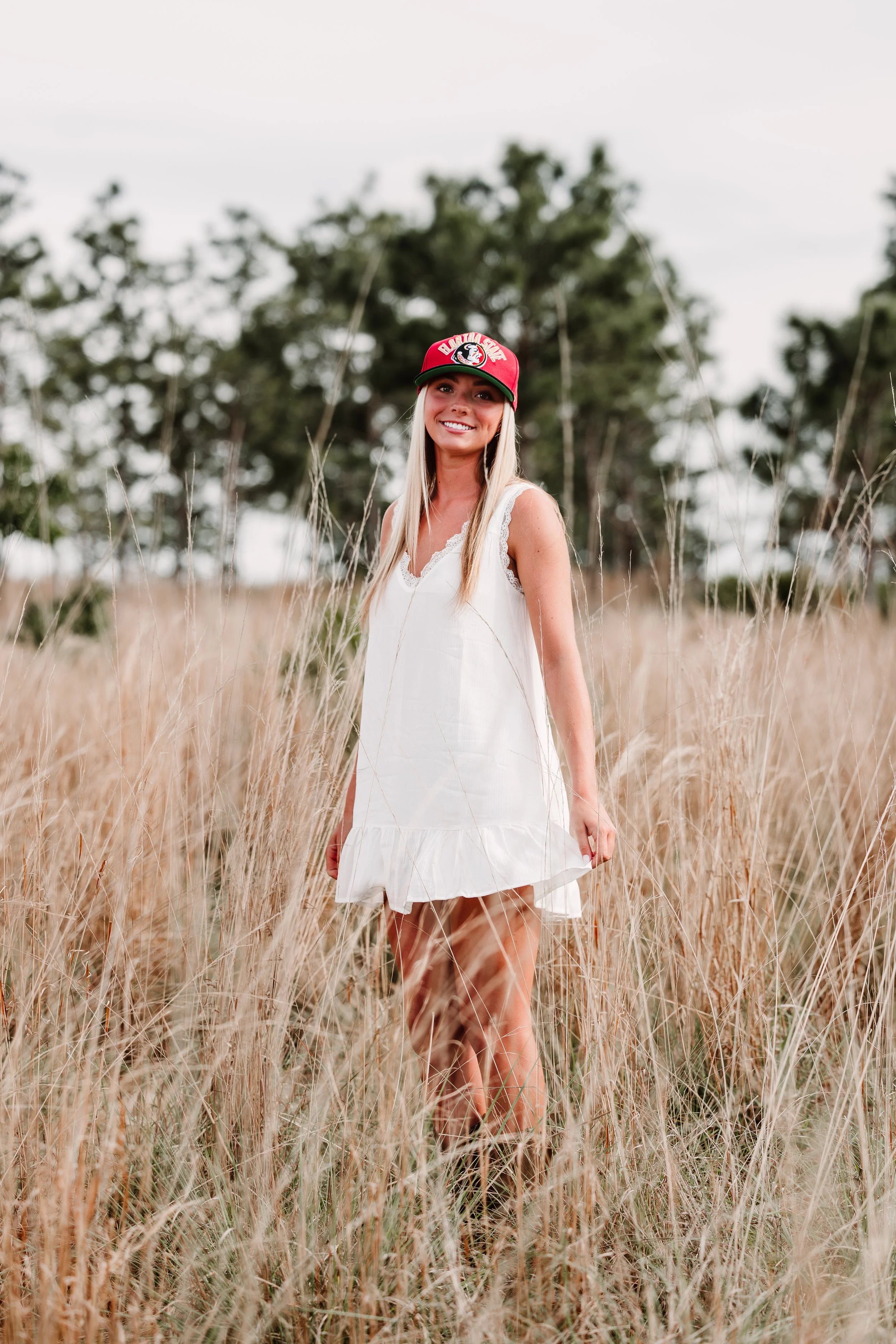 A woman with long blonde hair smiling and wearing a red baseball cap and a white dress stands in a field of tall, dry grass with trees in the background.