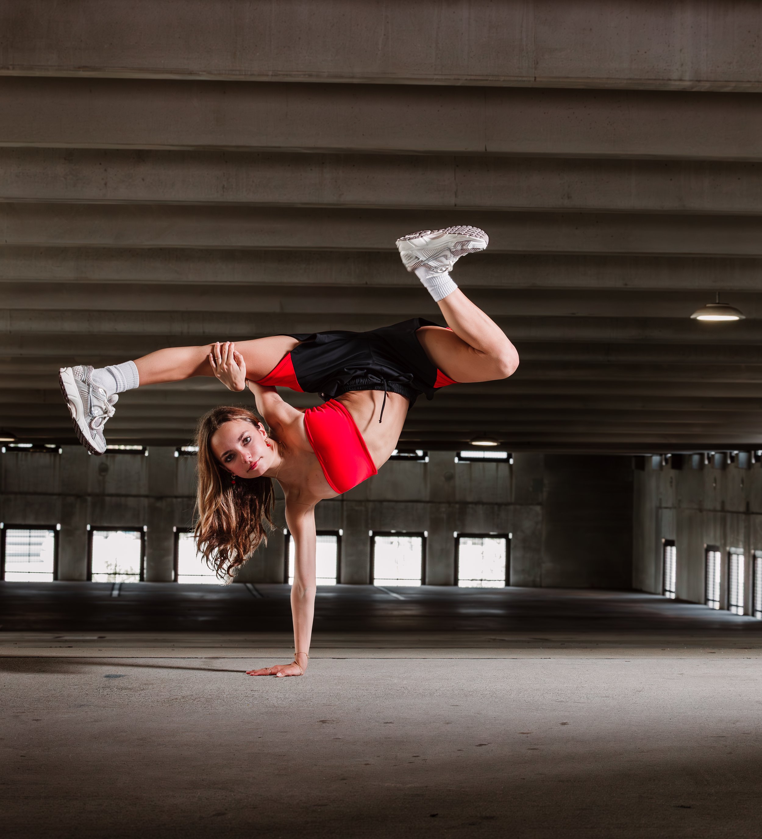 A woman in athletic clothing performing a one-handed handstand in an indoor parking garage.