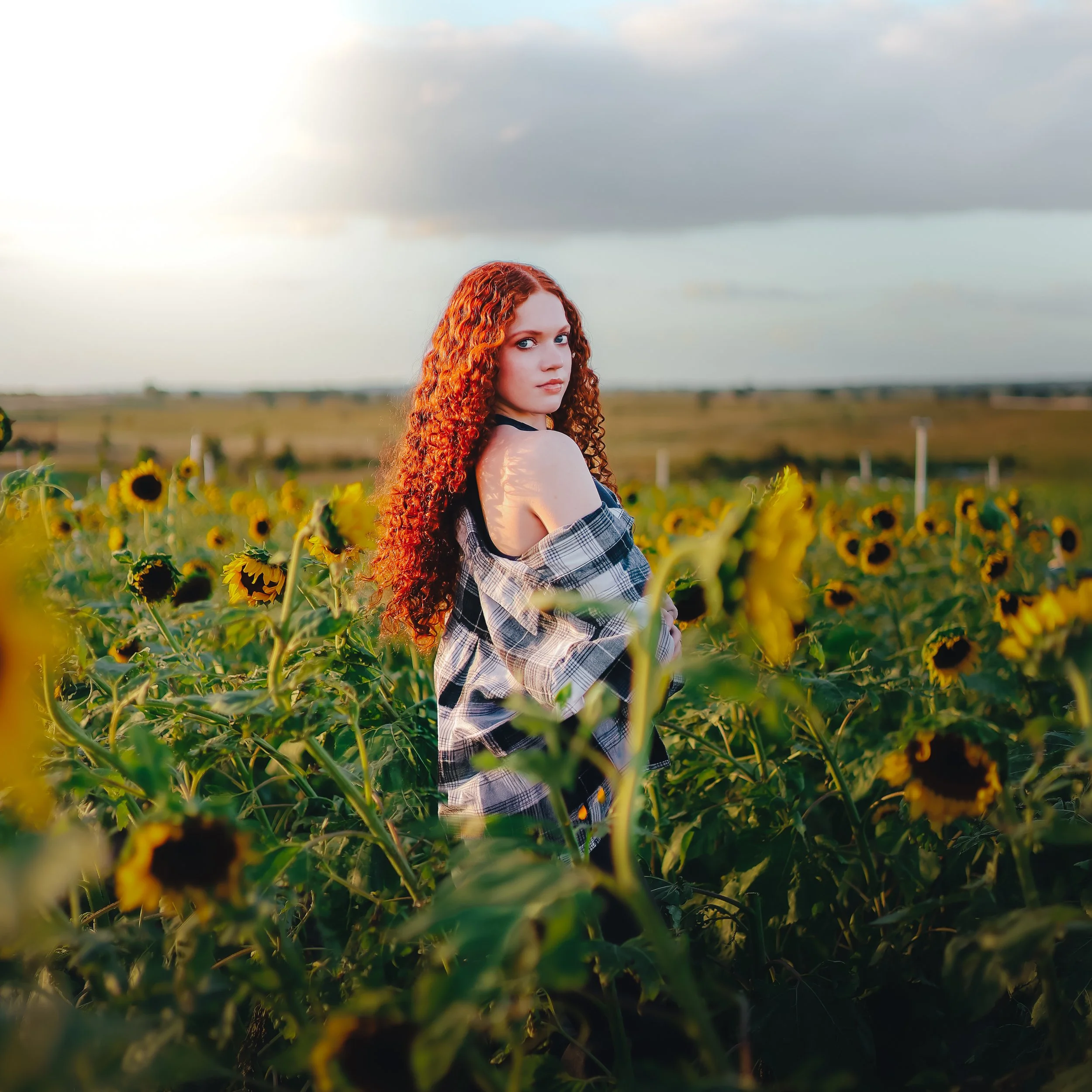 A woman with long, curly red hair standing in a sunflower field during the daytime.