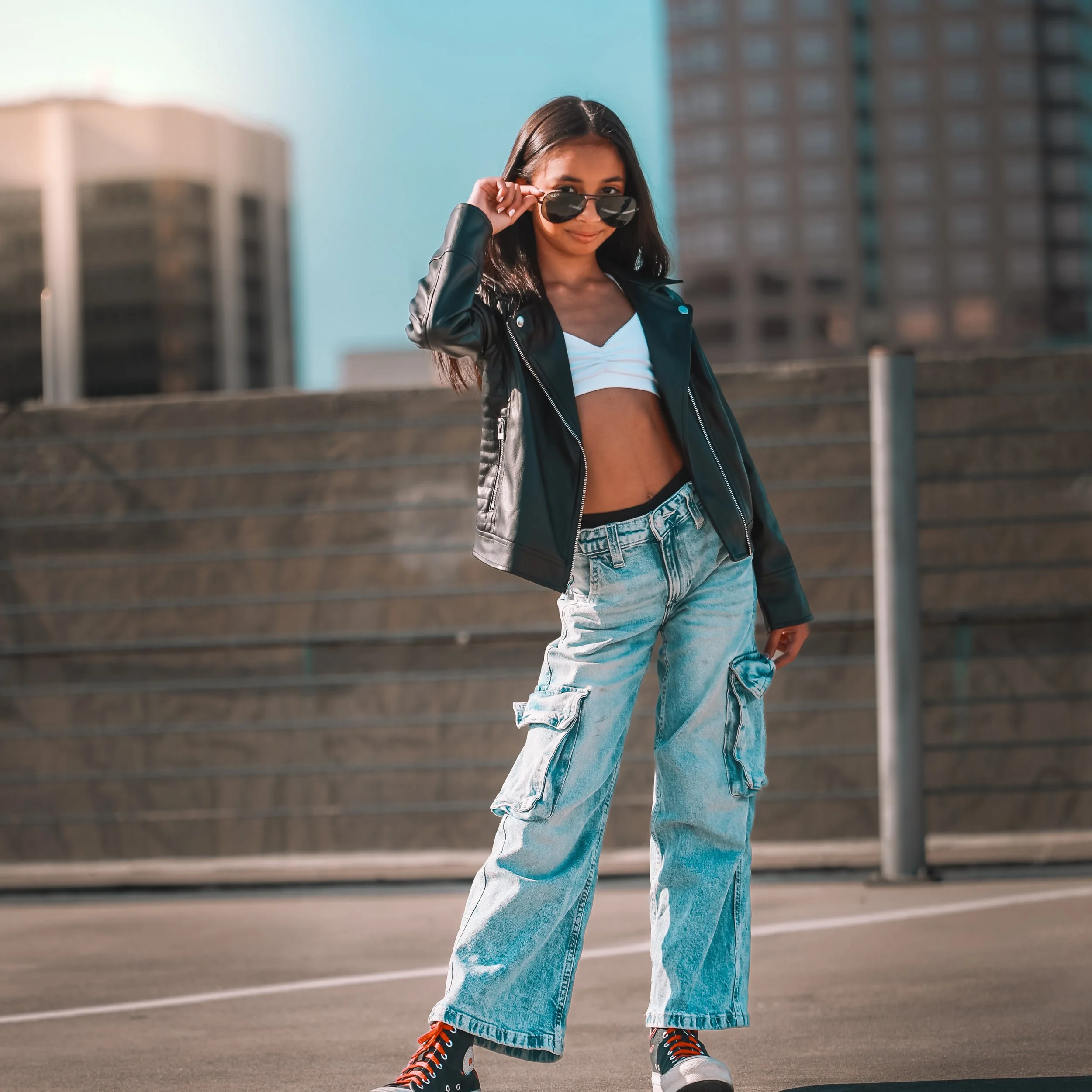 A young woman with long dark hair wearing sunglasses, a black leather jacket, a white crop top, baggy light wash jeans, and black sneakers with red laces, standing outdoors on a city street with tall buildings in the background.