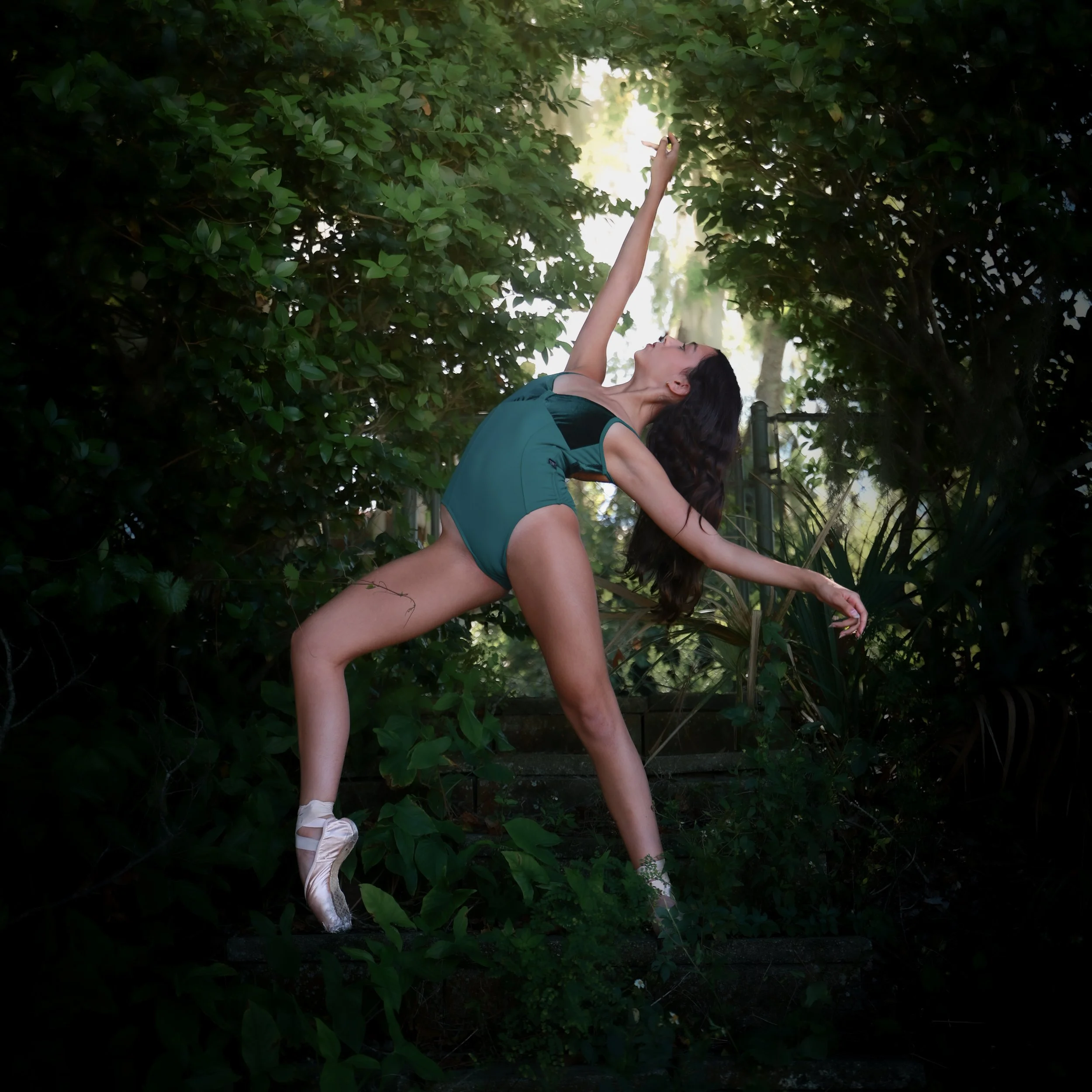 A woman in a teal shirt and ballet slippers poses gracefully among green bushes outdoors.