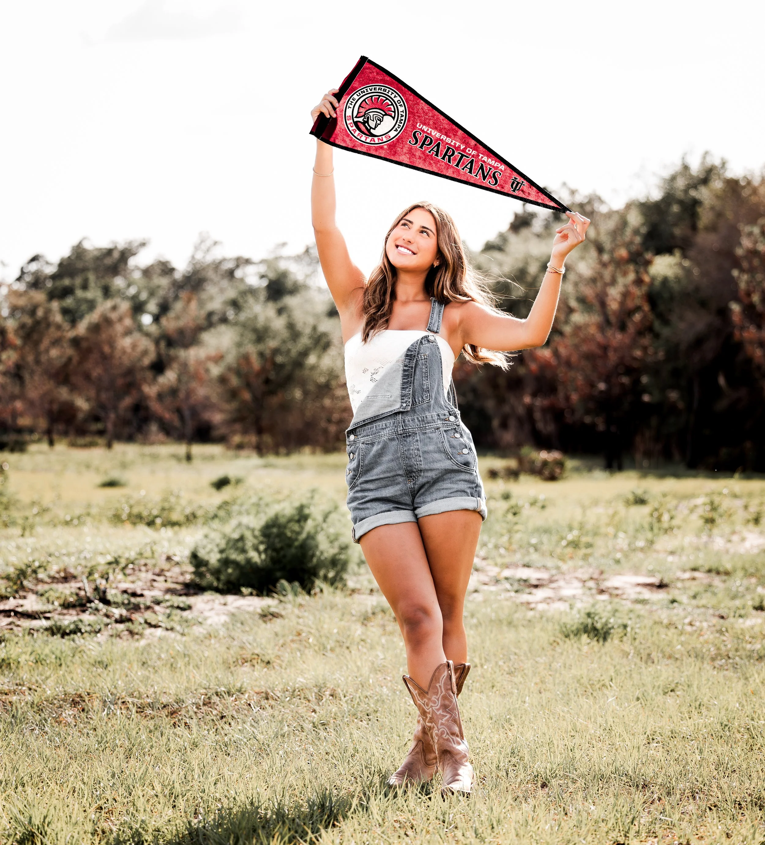 A young woman in denim shorts and cowboy boots holds a University of Tampa Spartans pennant in a grassy outdoor field with trees in the background, smiling.