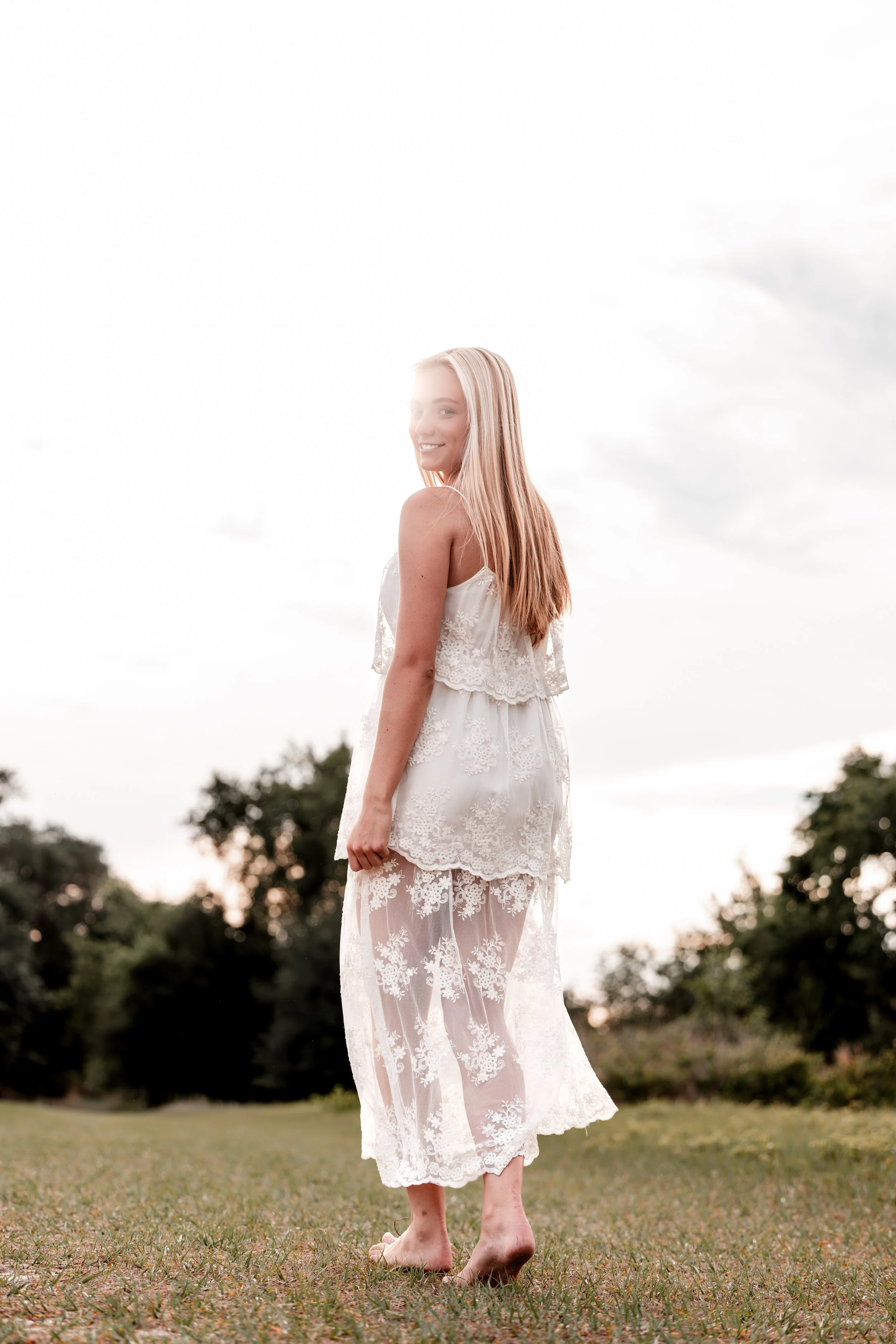 A young woman with long blonde hair wearing a white lace dress, standing outdoors on grass with trees in the background. She is smiling and looking over her shoulder, with her back to the camera.