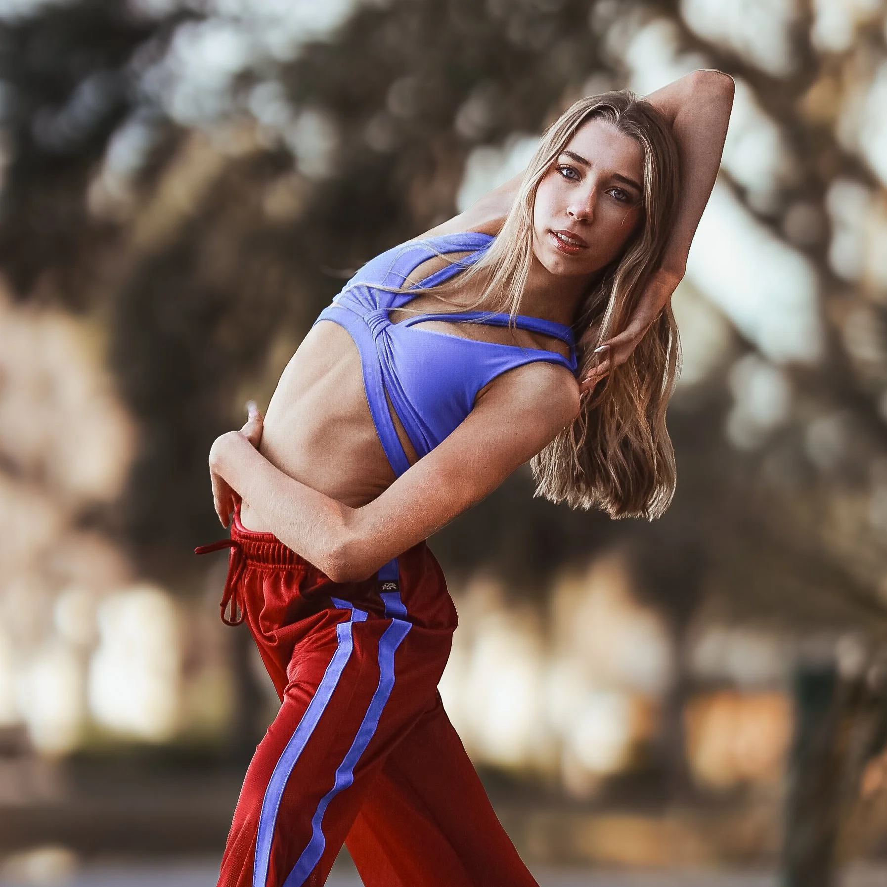 A young woman with long blonde hair poses outdoors, wearing a blue crop top and red athletic pants with blue stripes, with a blurred natural background.