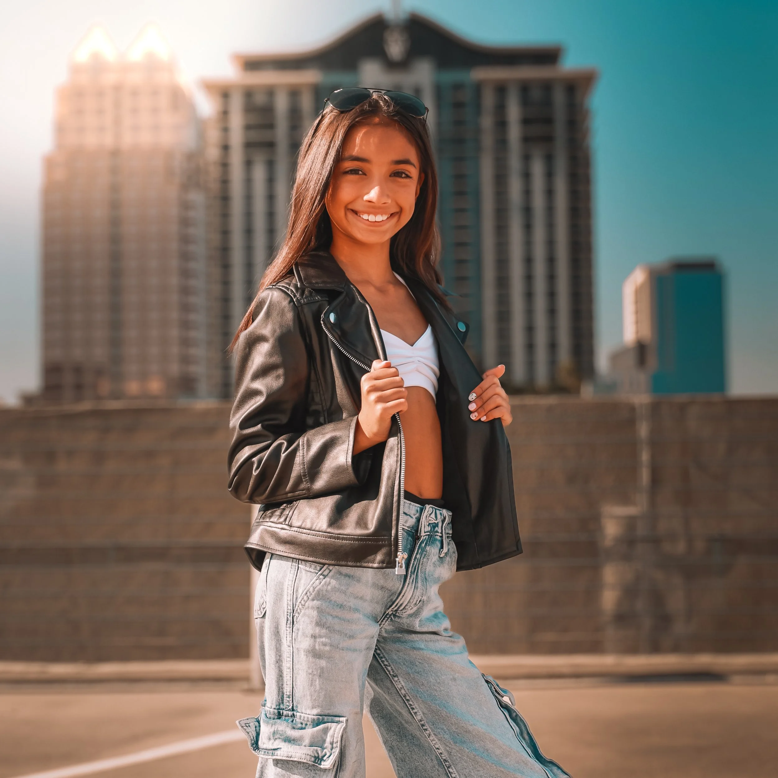Portrait of a smiling young woman with long brown hair wearing sunglasses on her head, a black leather jacket, and distressed jeans, standing outdoors in front of tall buildings during daytime.