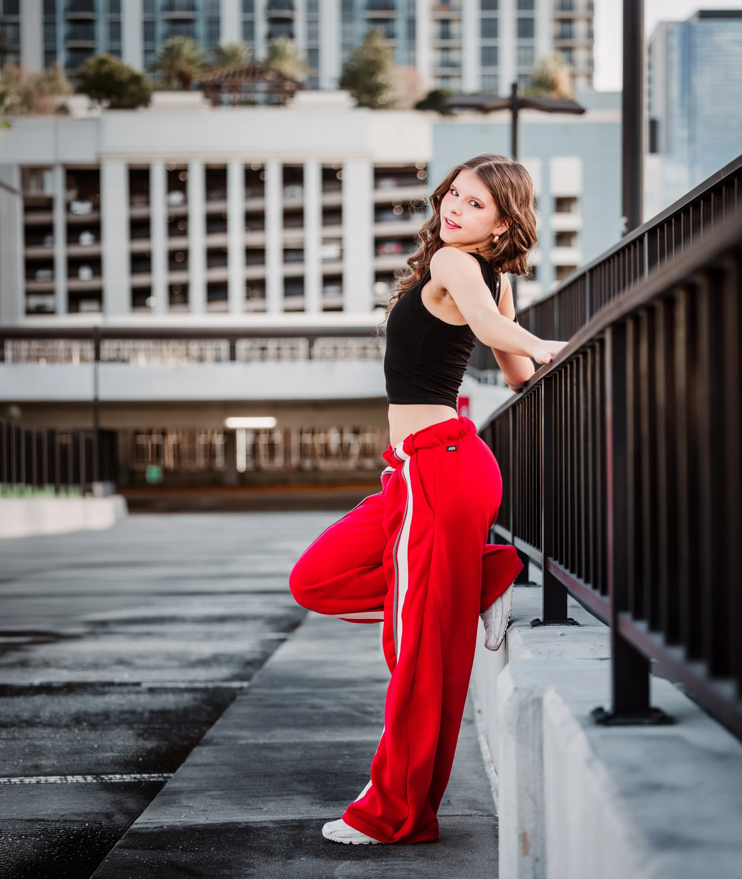 A young woman with long wavy brown hair wearing a black crop top and red sweatpants with white sneakers is kneeling on one knee on a city rooftop parking lot with a black metal railing, with tall modern buildings and palm trees in the background.