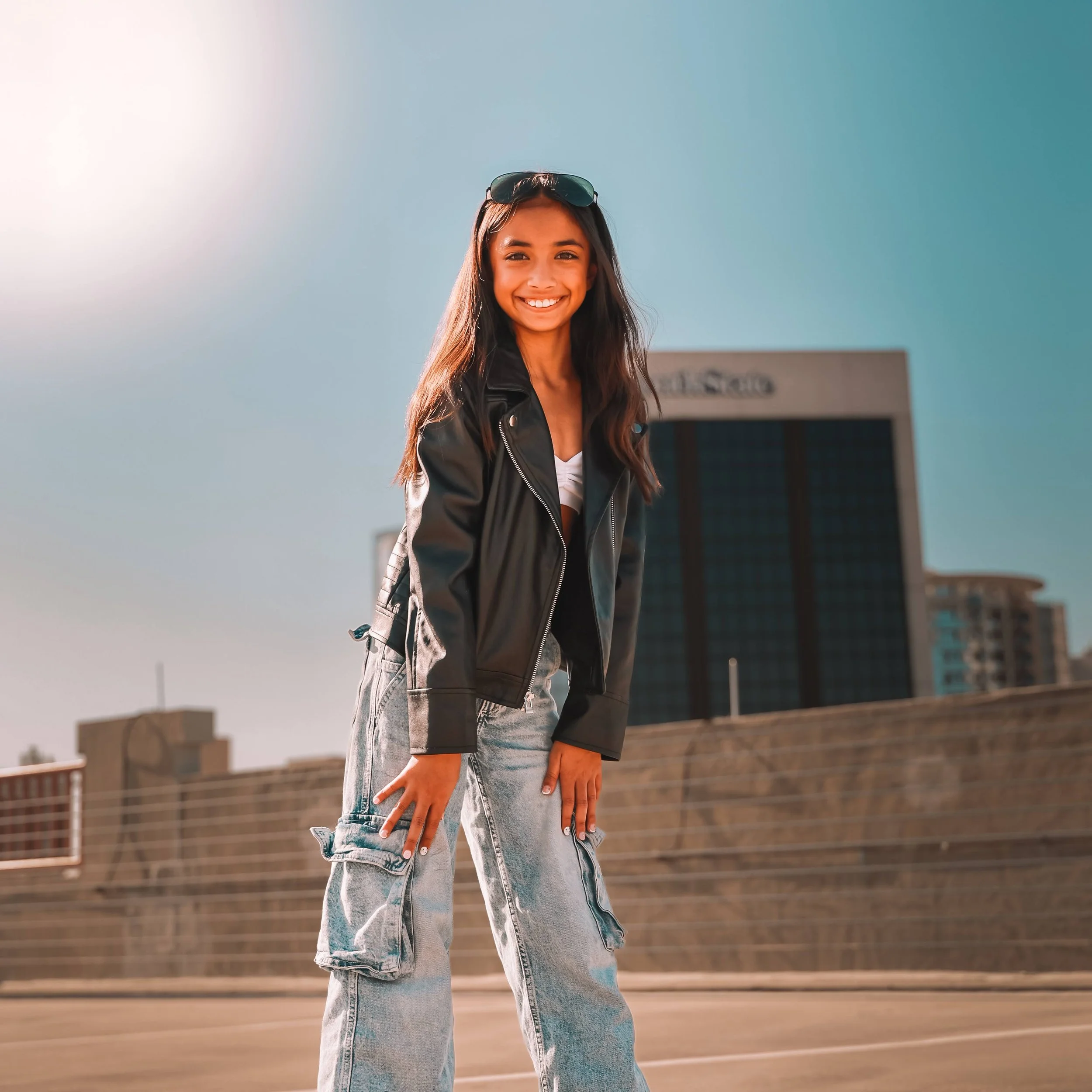 A young woman with long brown hair and a big smile standing outdoors in front of a clear blue sky and a cityscape. Lifestyle portrait photography in Orlando FL.