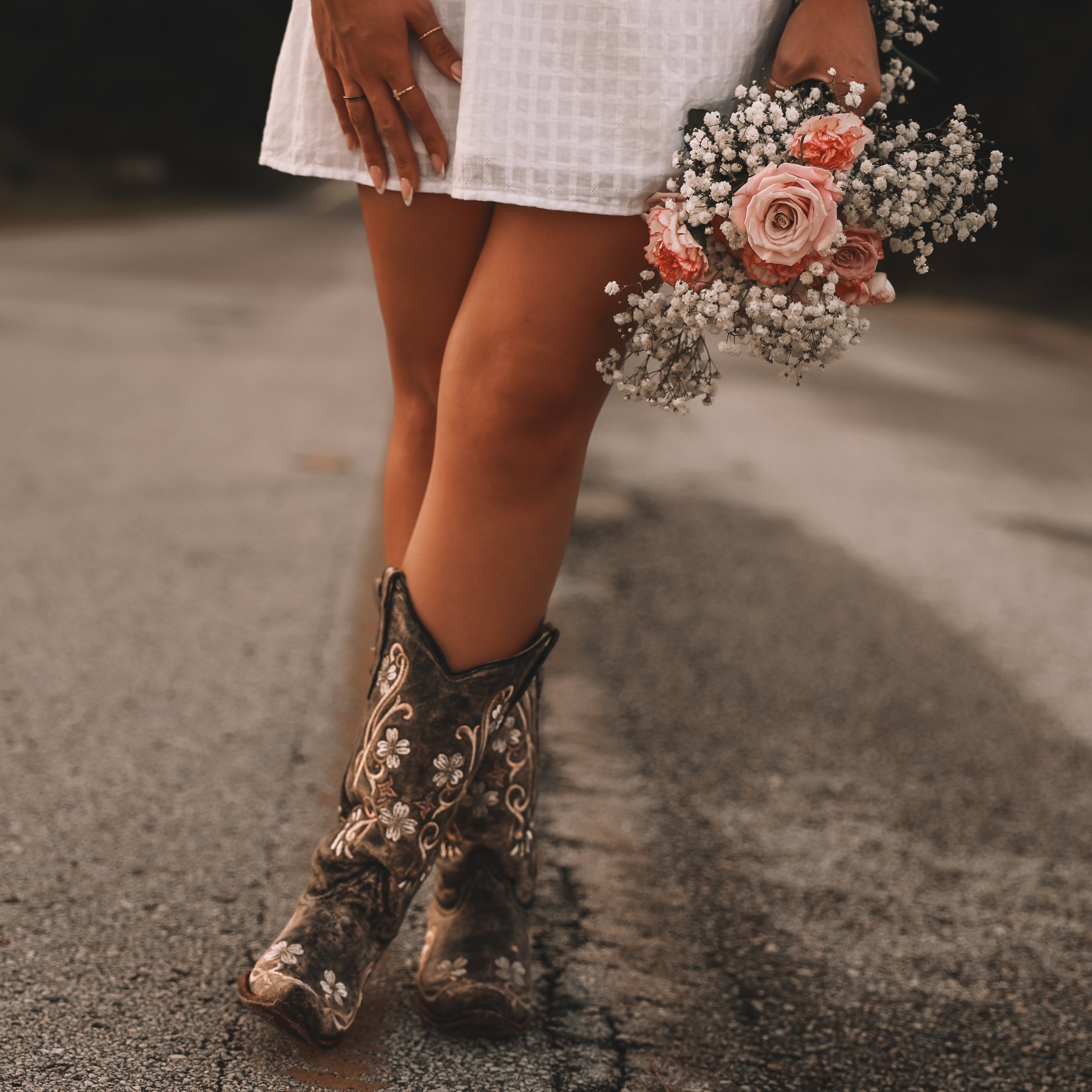 Woman holding a bouquet of pink and white flowers while wearing decorative cowboy boots, standing on a textured outdoor surface.