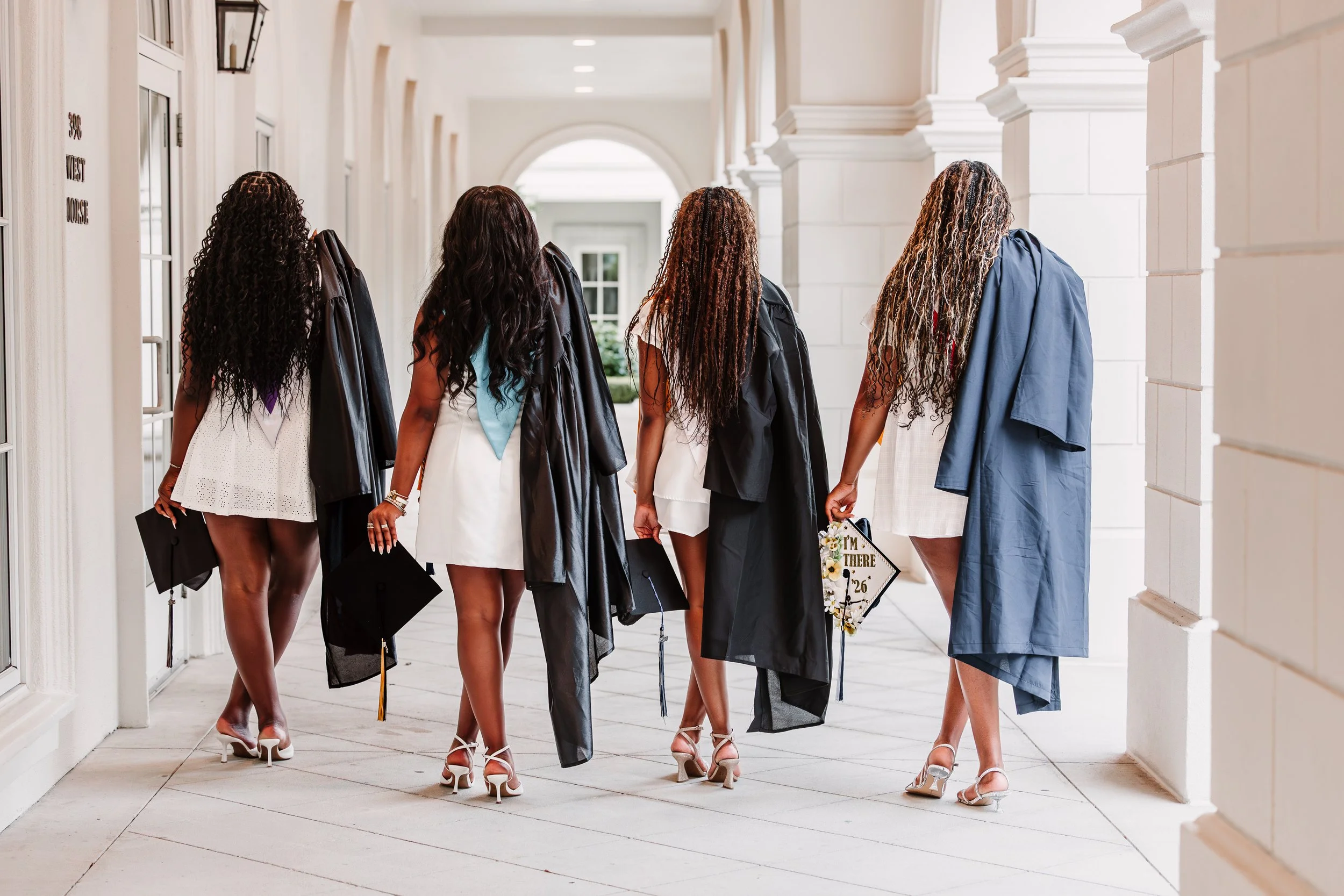 Four women wearing graduation gowns and caps walk down a bright, white corridor outside a building, holding themselves with confidence. They have curly and styled hair, and some hold flowers or signs.